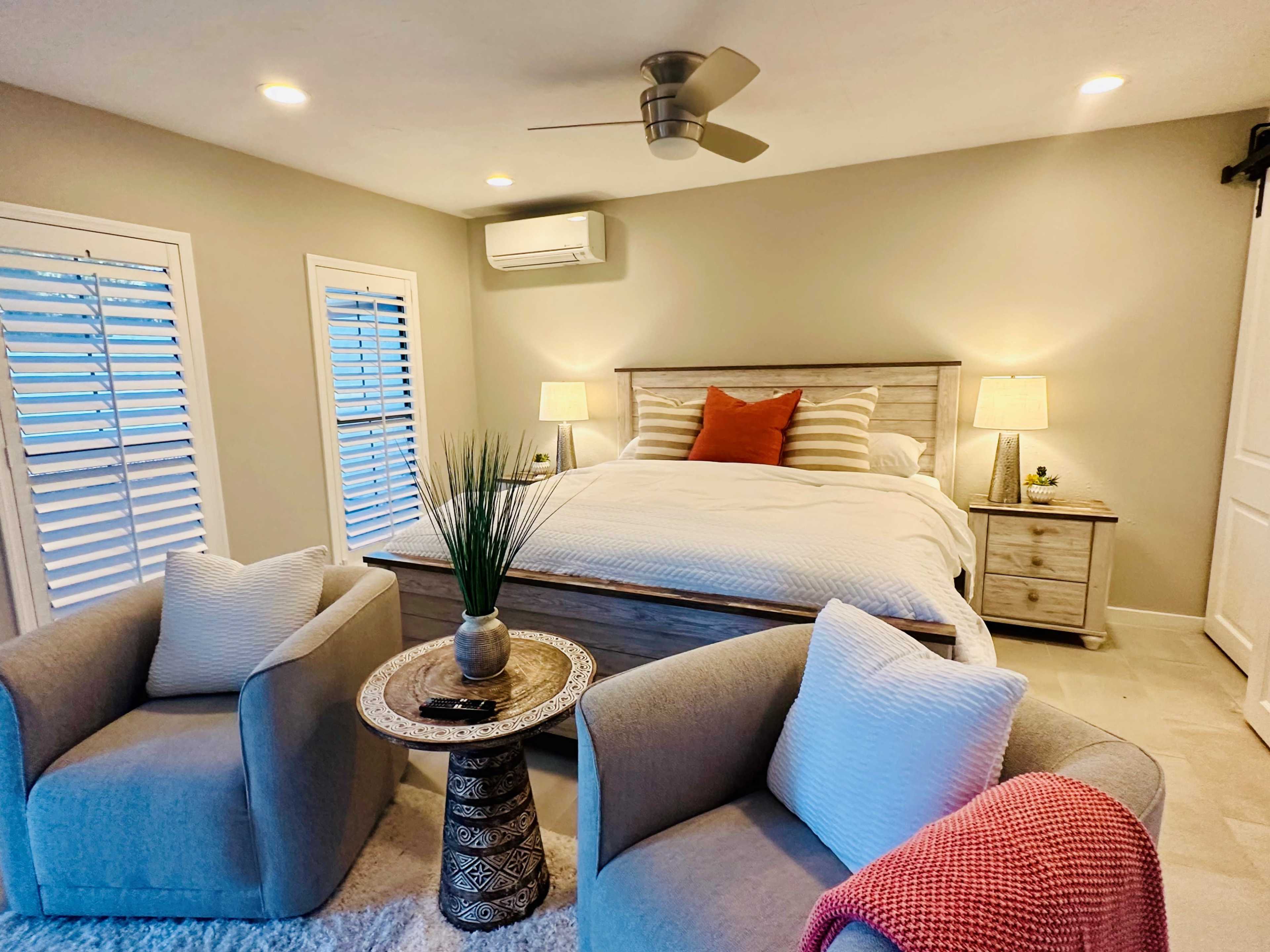 A cozy bedroom features a double bed with striped linens, two bedside lamps, and a seating area with two gray chairs and a decorative table.