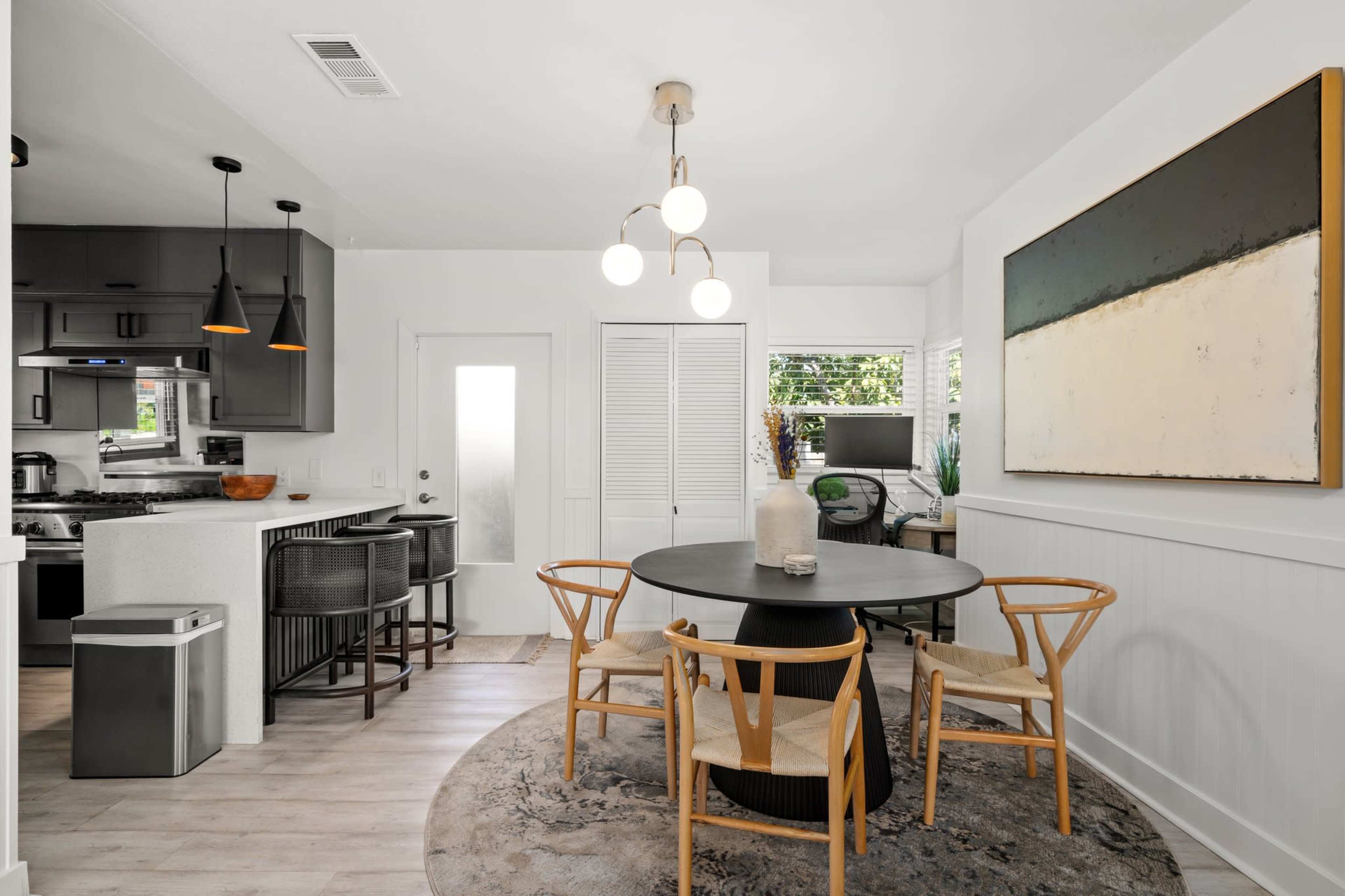 A dining area features a round table surrounded by four chairs, with a modern kitchen visible in the background and a large abstract painting on the wall.