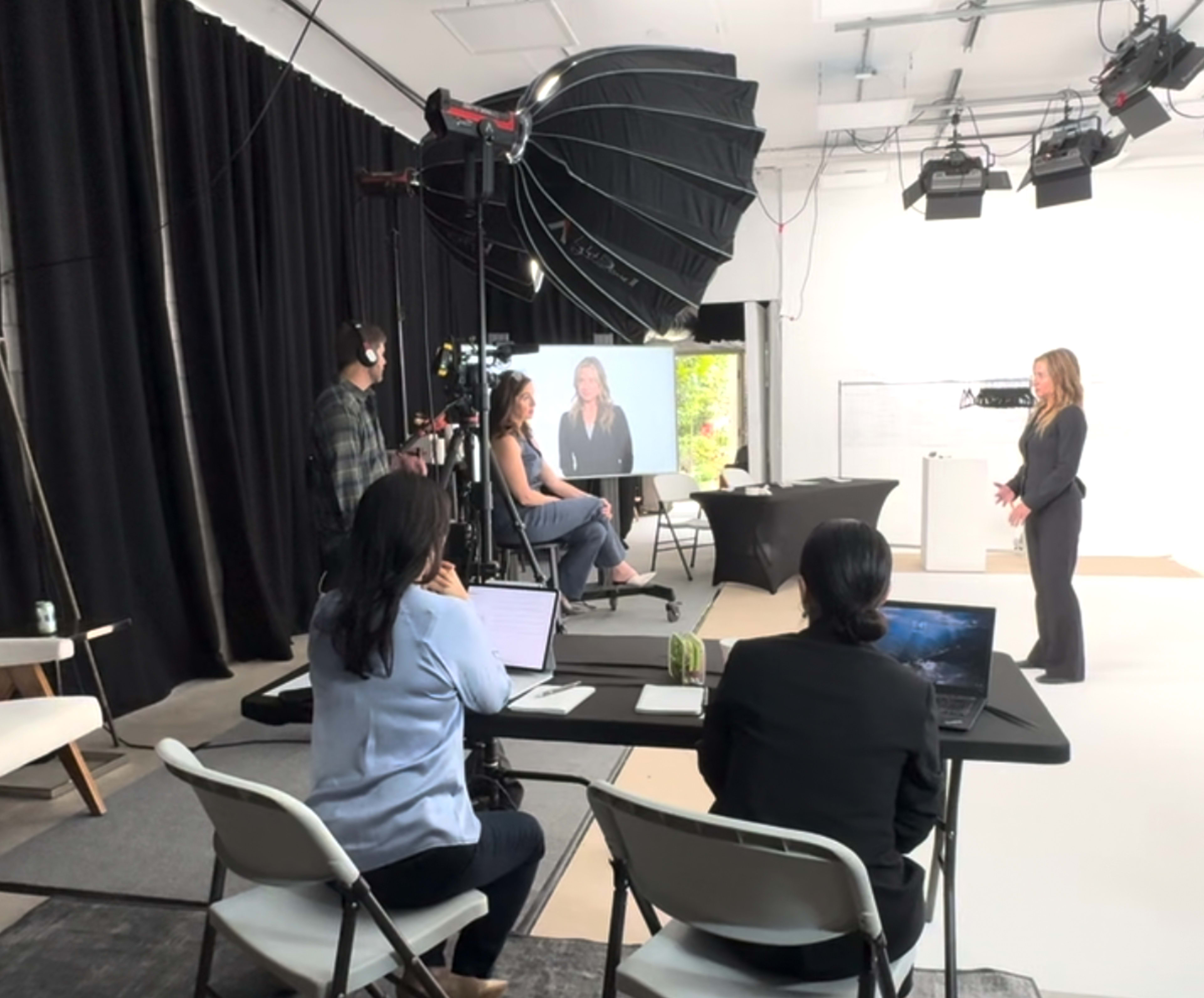 A group of people is set up for a video production with a camera and lights, while two women are visible on screen in a studio environment.