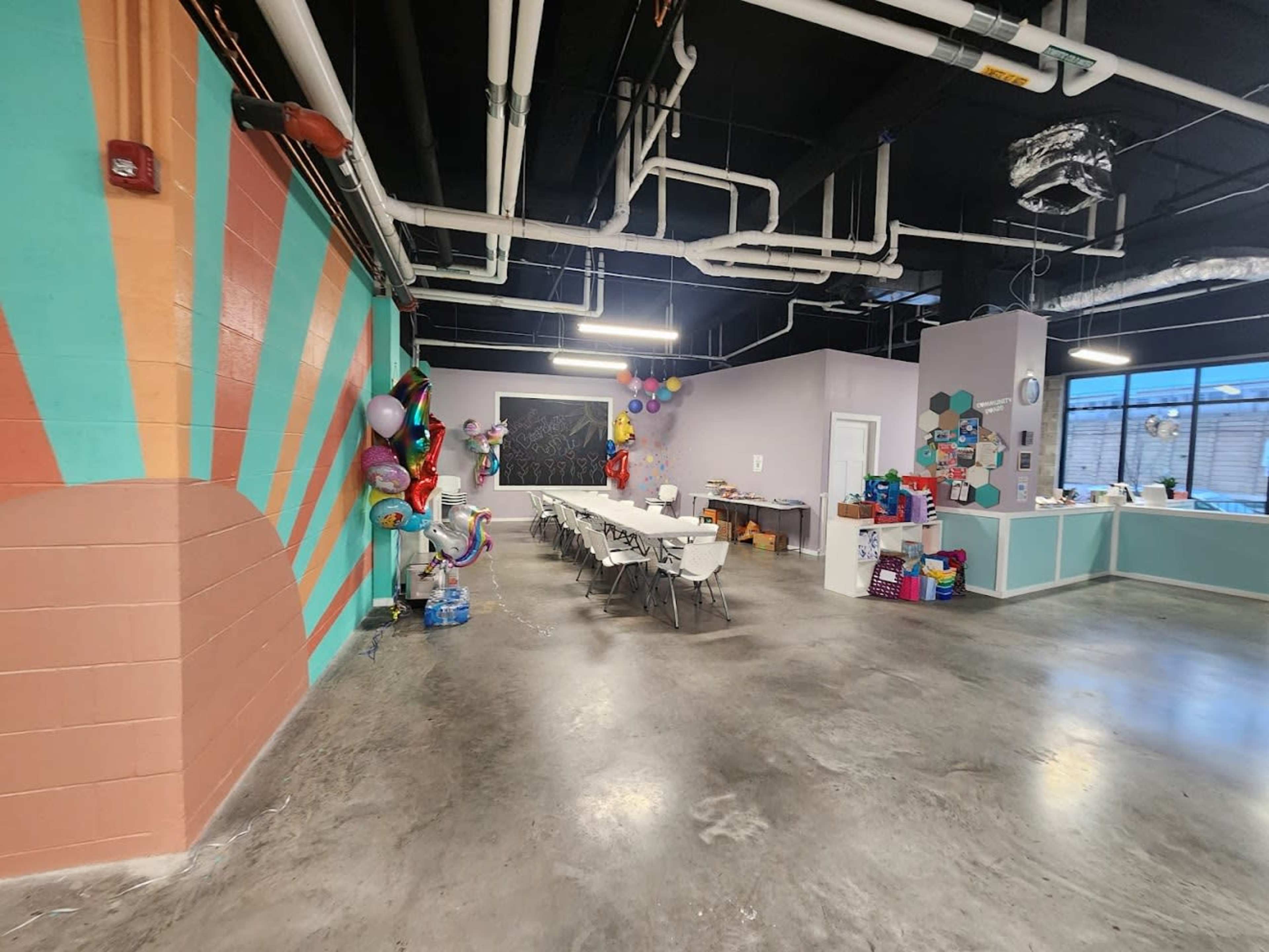 An indoor event space featuring a long table with chairs, colorful wall decorations, and party balloons in one corner.