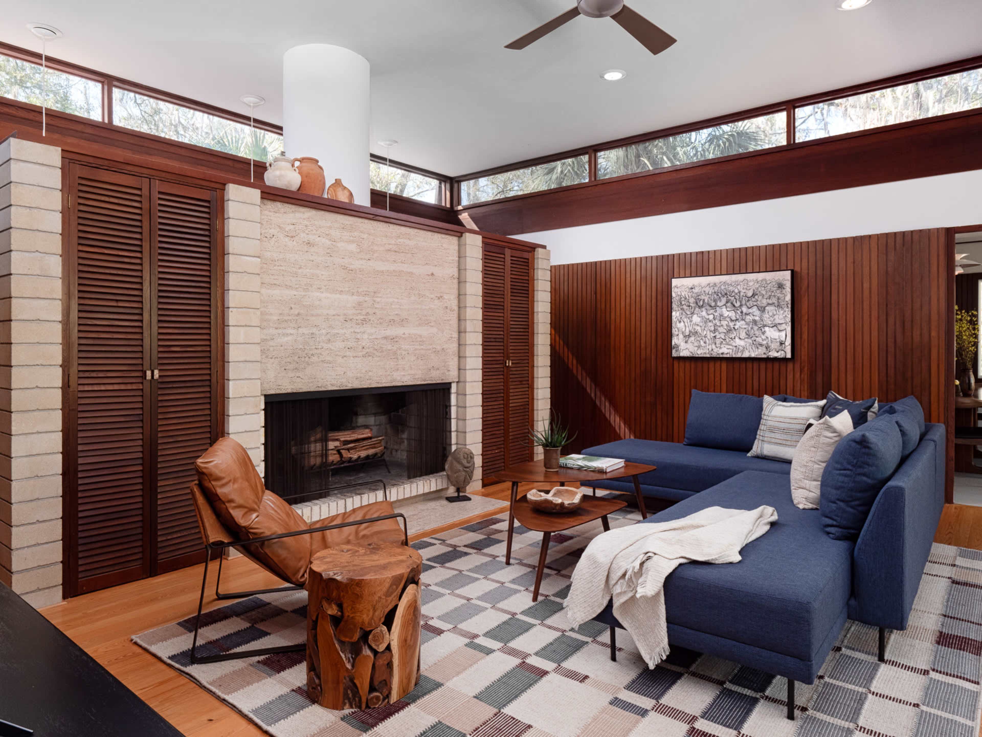 The image shows a modern living room with a large blue sectional sofa, a circular wooden coffee table, a stone fireplace, and wooden cabinetry.