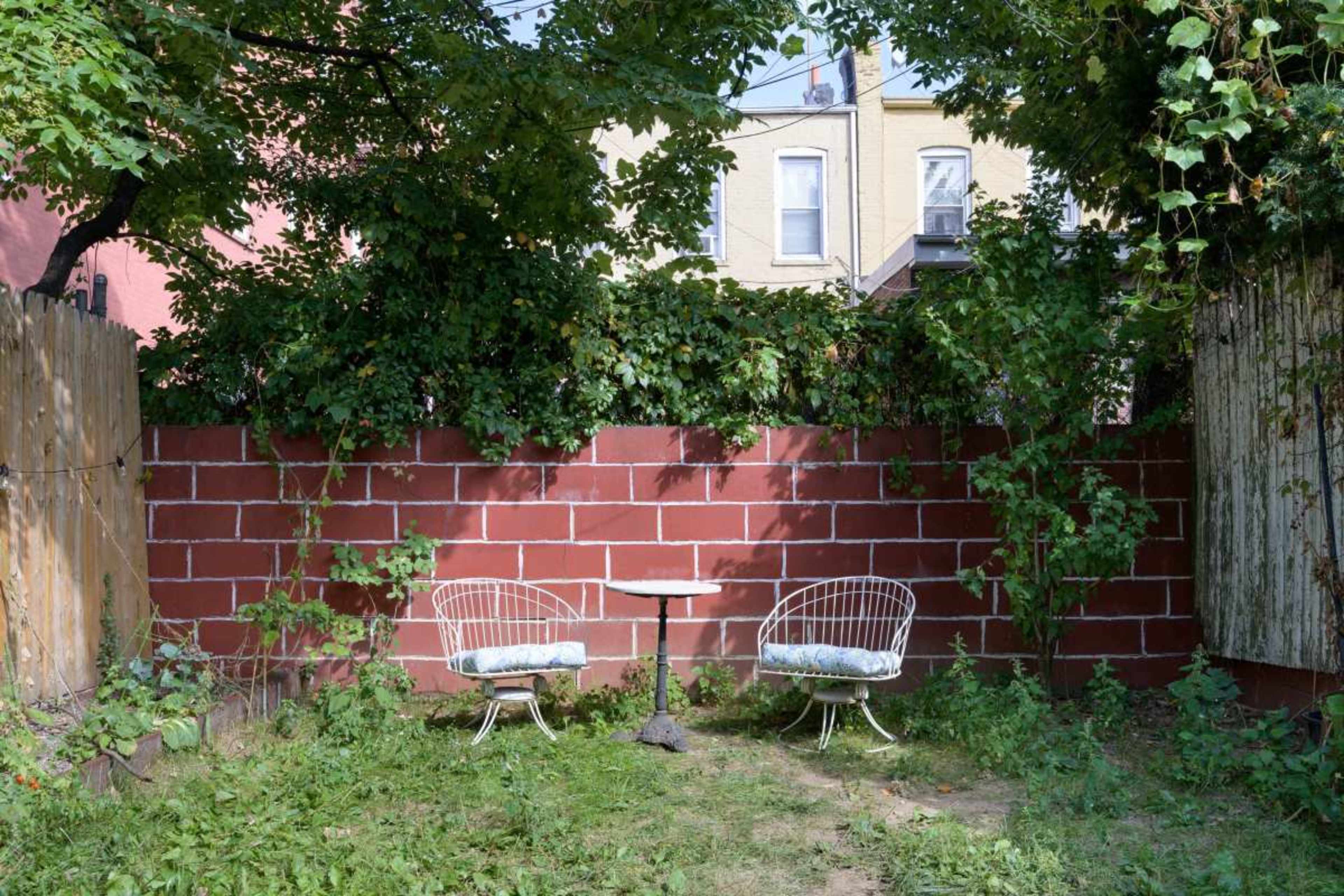 A small, enclosed garden featuring two white metal chairs and a table against a red brick wall, surrounded by greenery.