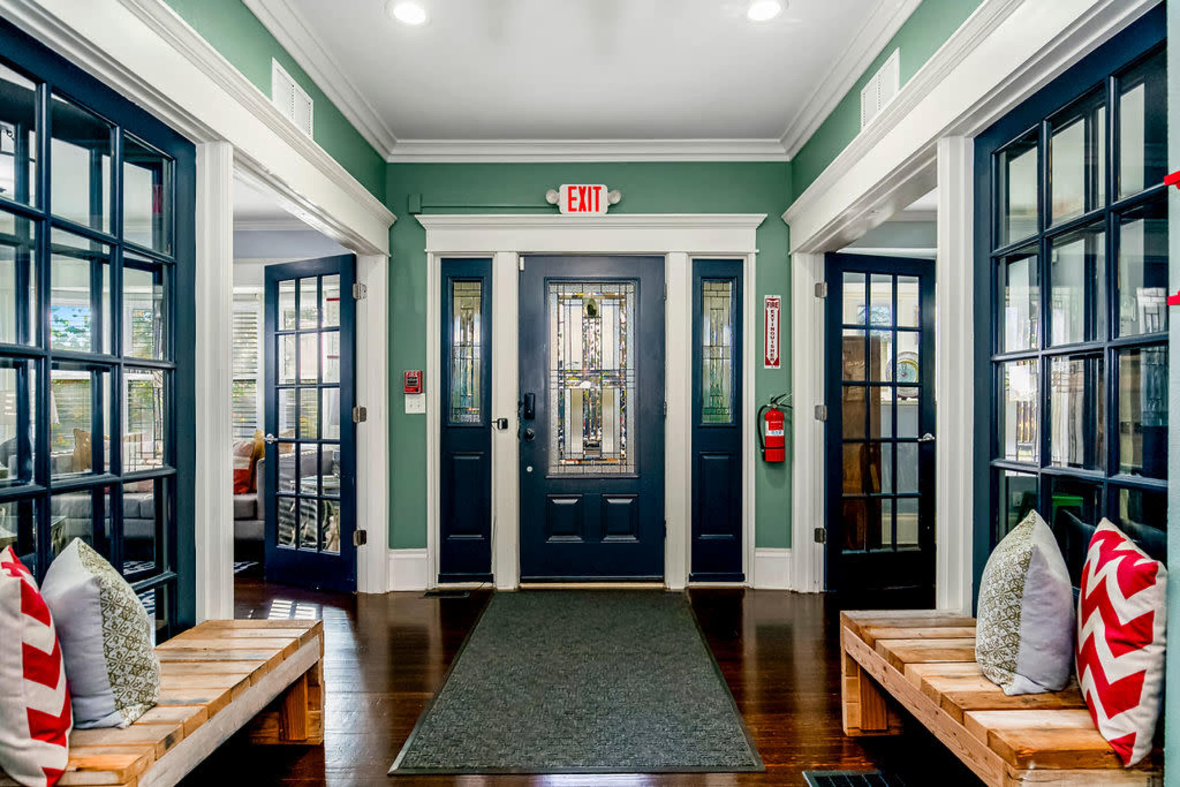 The image shows a well-lit hallway with a decorative front door, flanked by two sets of glass-paneled doors and wooden benches on either side.