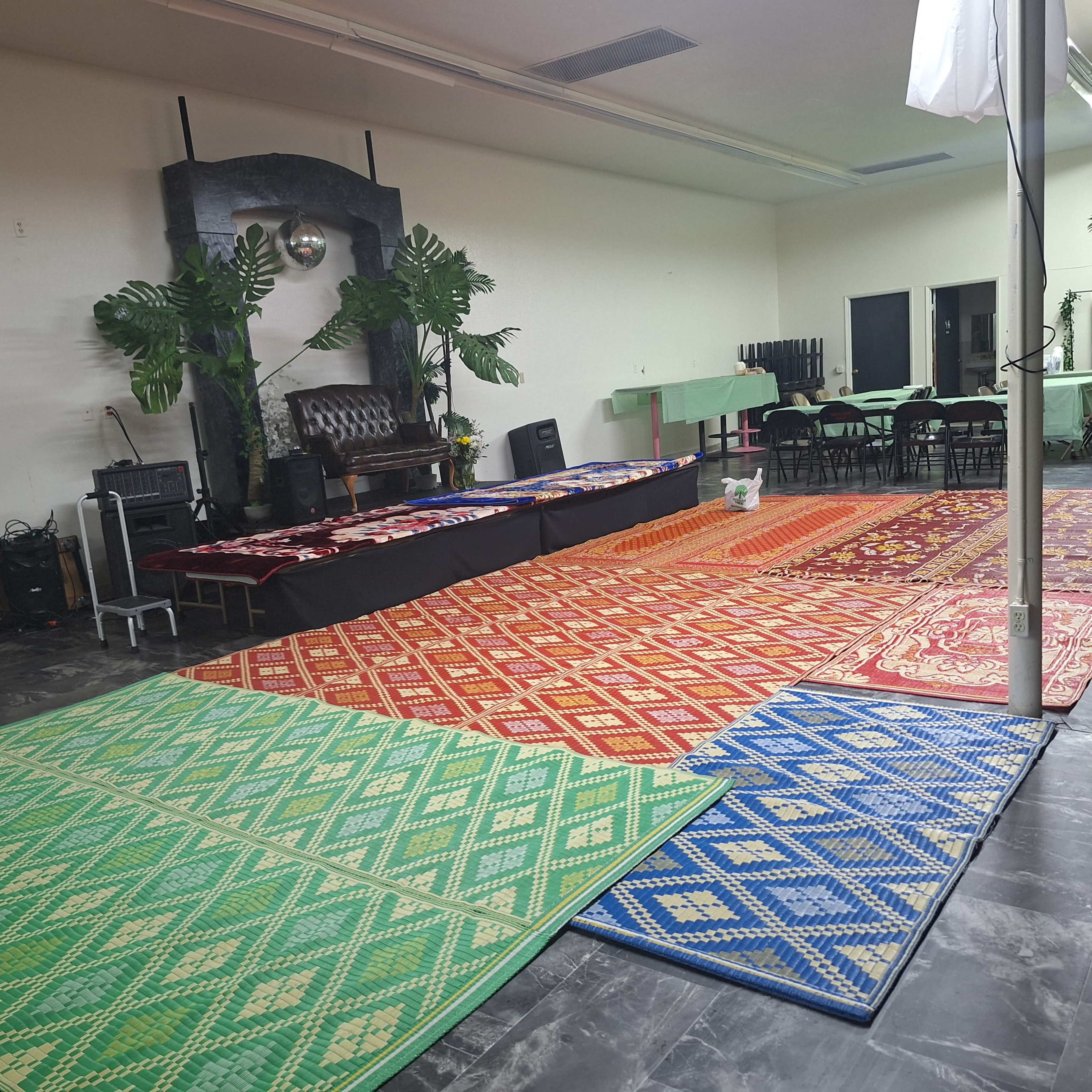The image shows a hall arranged with colorful patterned rugs covering the floor, while tables are set in the background.