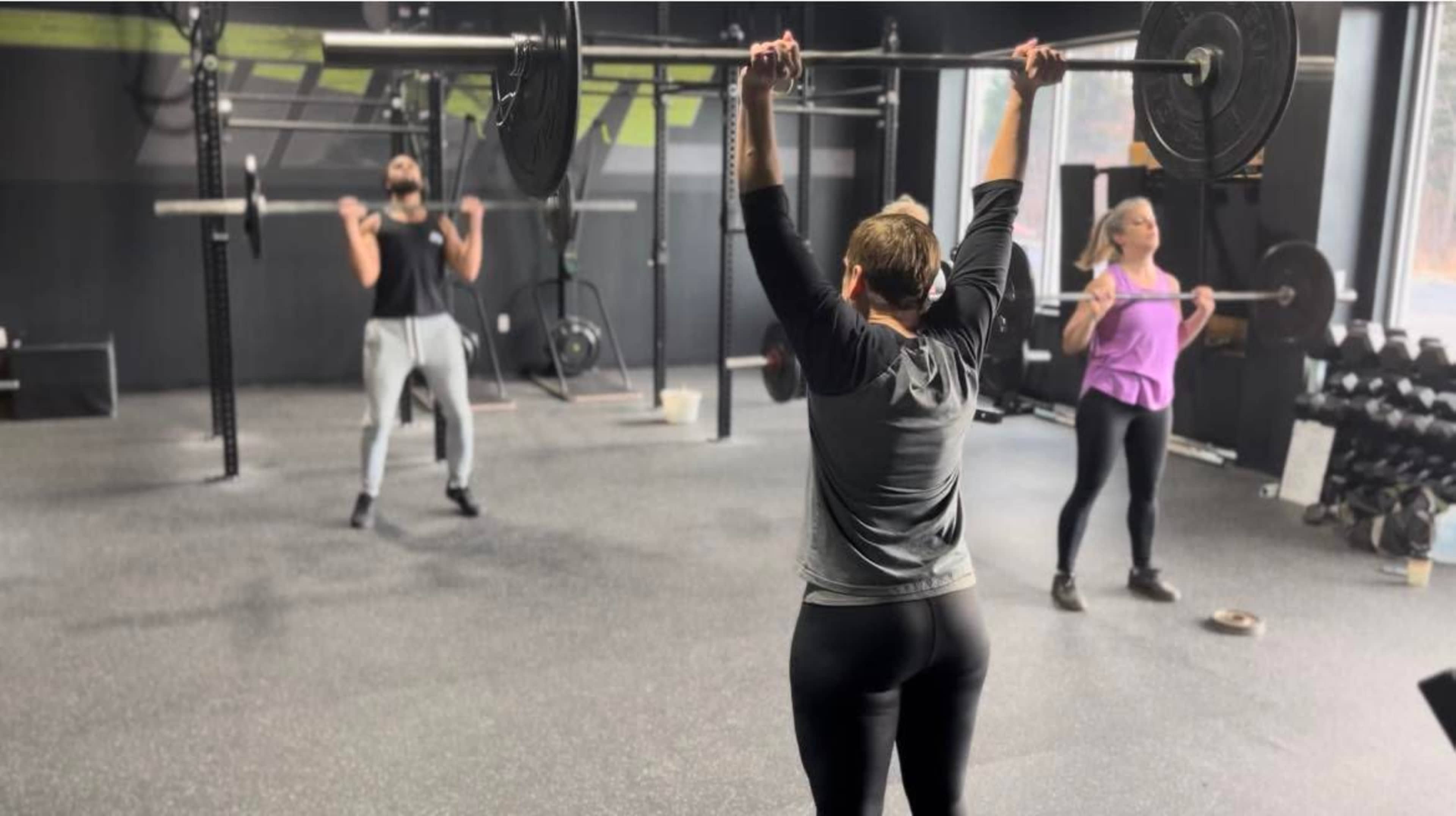 Three individuals are exercising in a gym, with one person lifting a barbell overhead while the others perform different weightlifting movements.