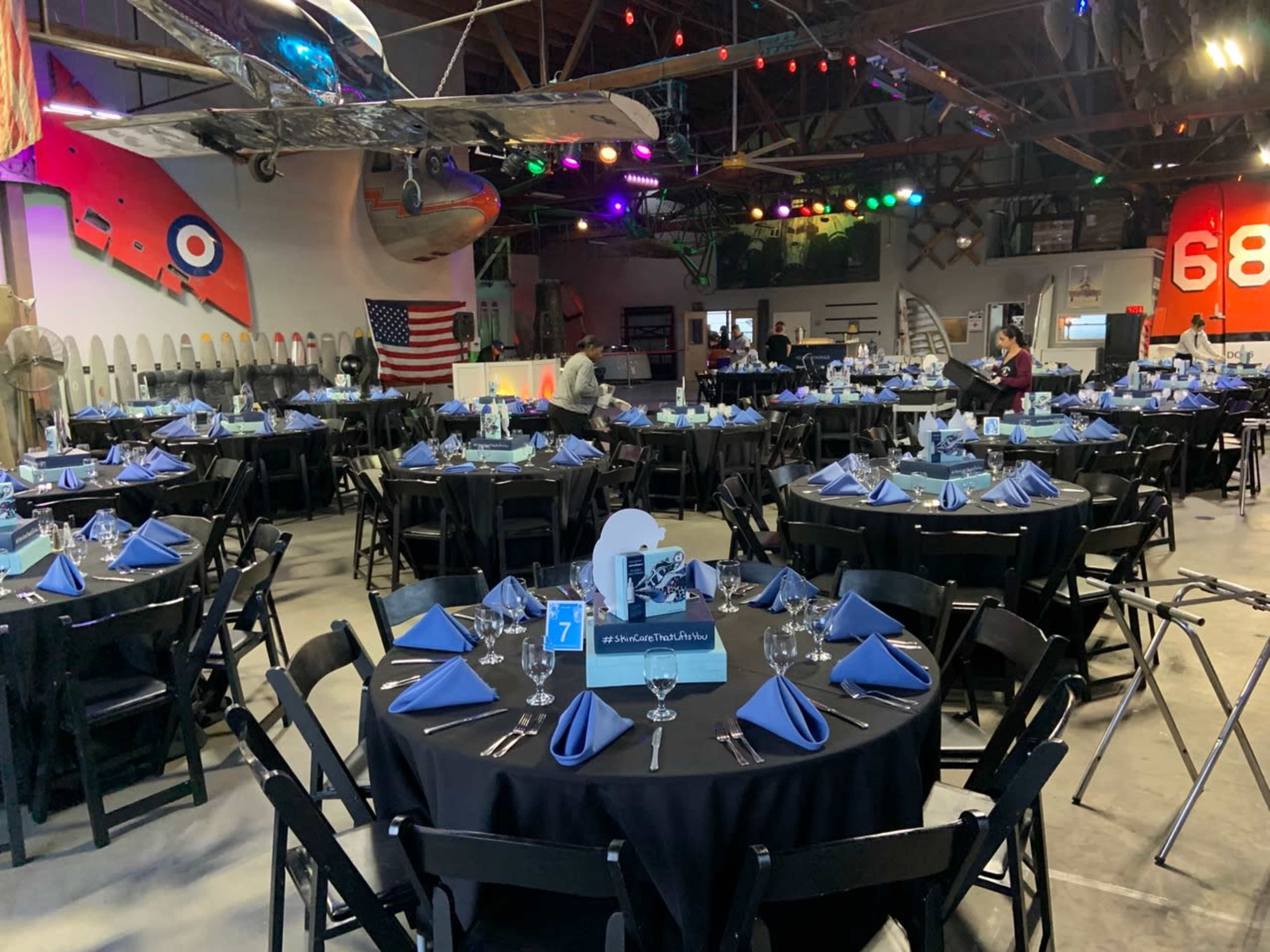 The image shows a banquet hall set up with round tables covered in black tablecloths, each adorned with blue napkins and glassware, under colorful lights and vintage aircraft decor.