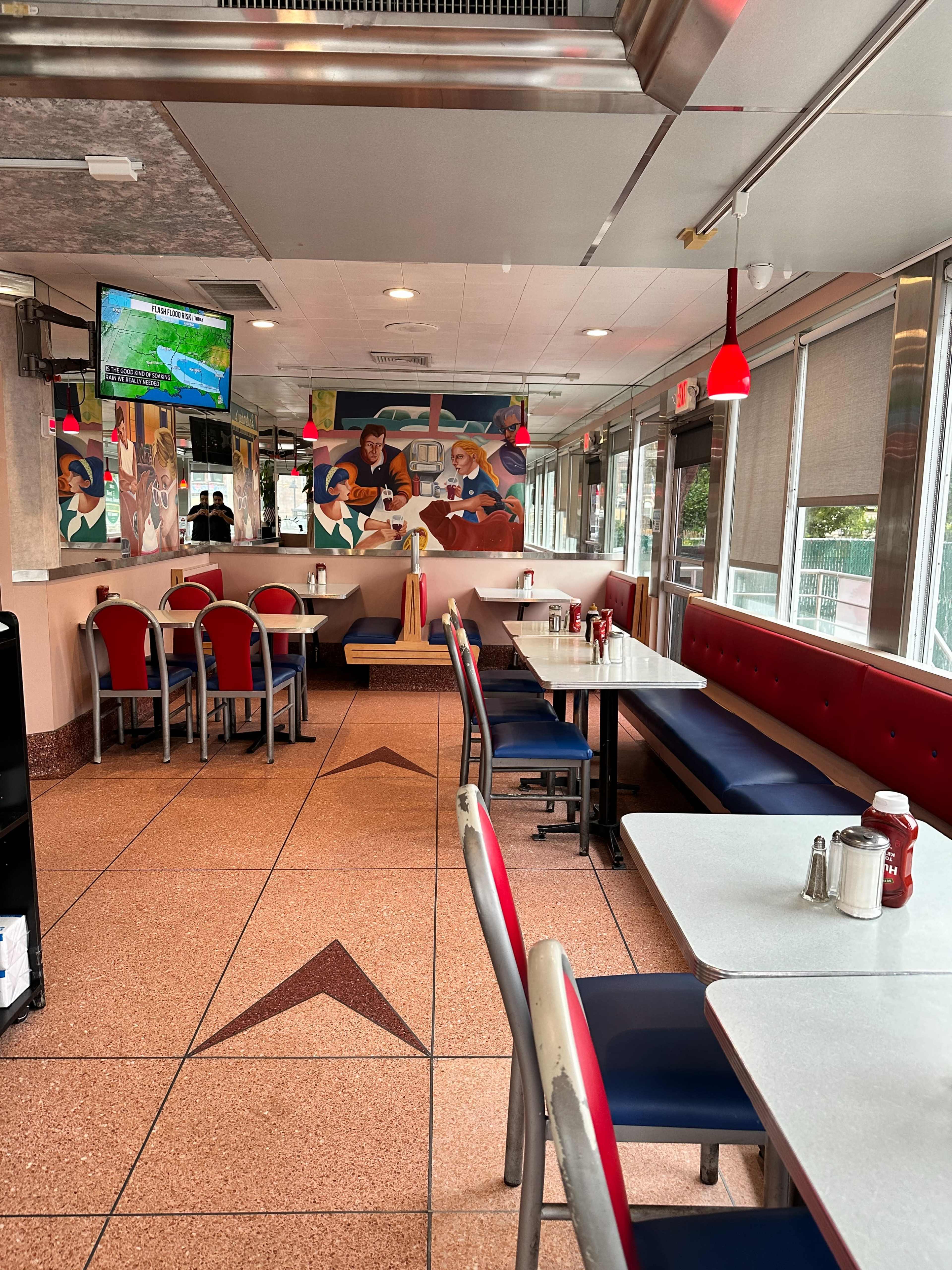The interior of a diner features several empty tables, red accents, and a mural on the wall depicting people dining.