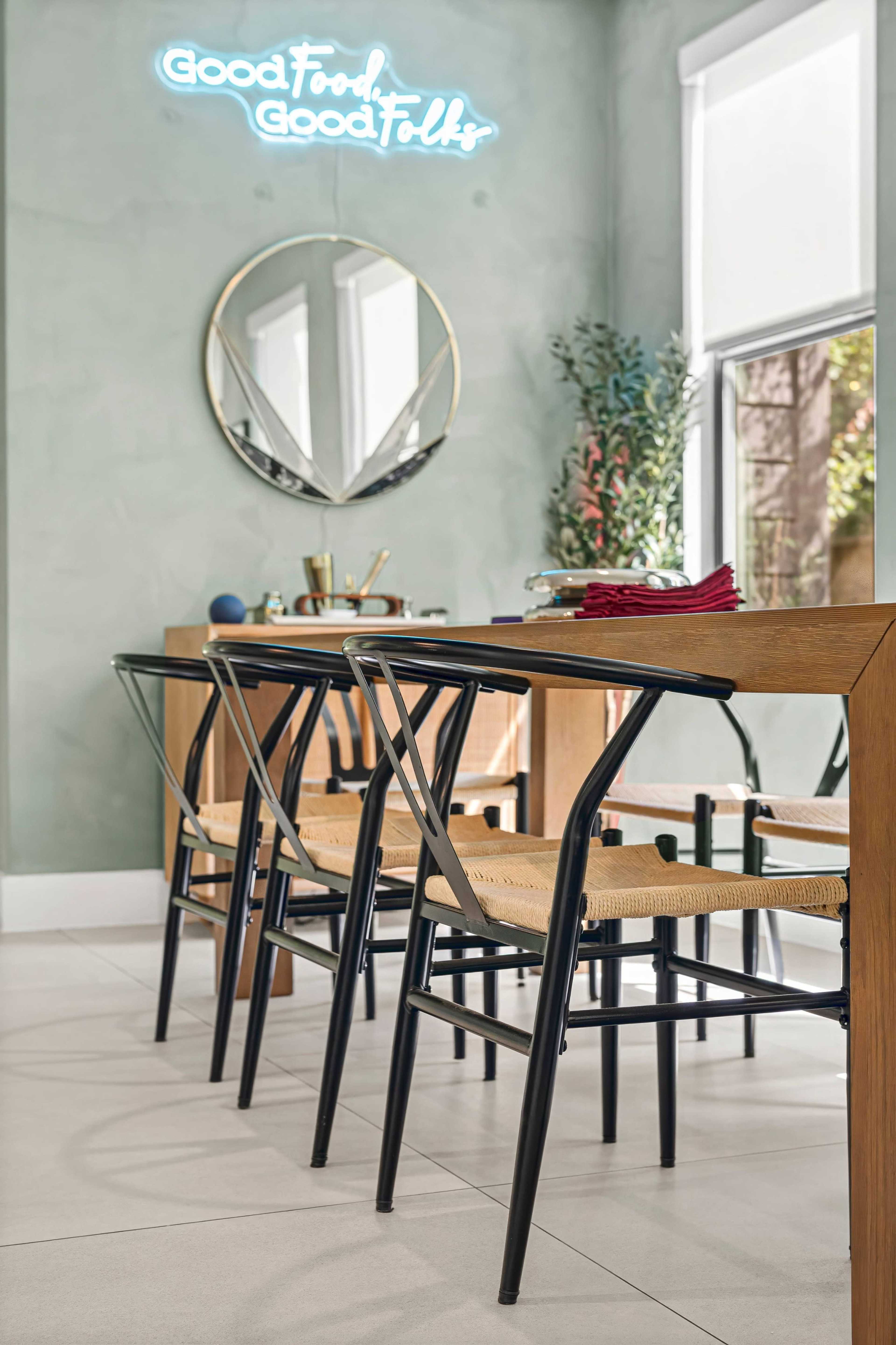 A simple dining area features a wooden table with several black and natural woven chairs, accompanied by a round mirror and a neon sign that reads "Good Food Good Folks."