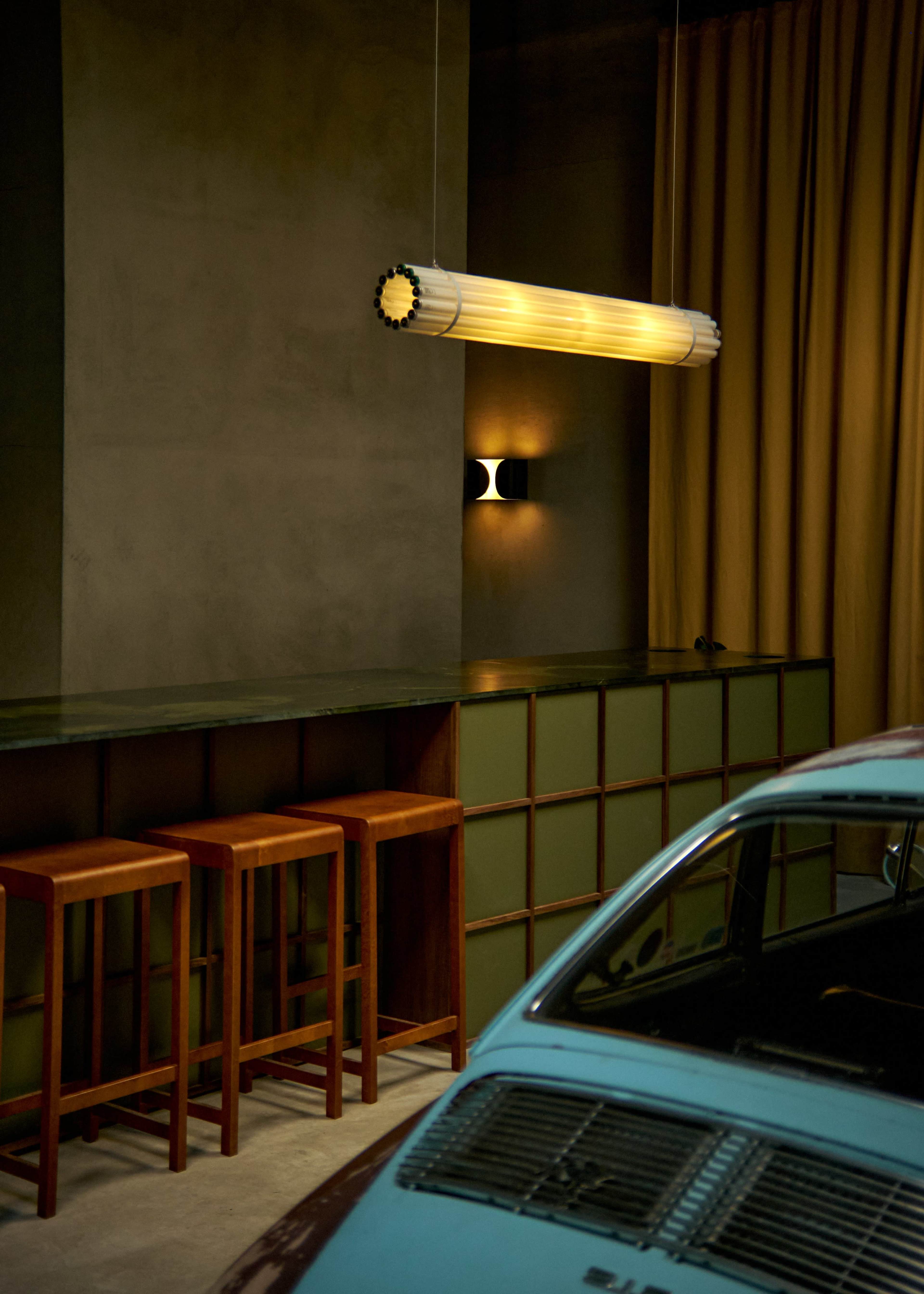 The image shows a dimly lit room featuring a retro car parked near a wooden bar with three tall stools, illuminated by a long hanging light fixture.