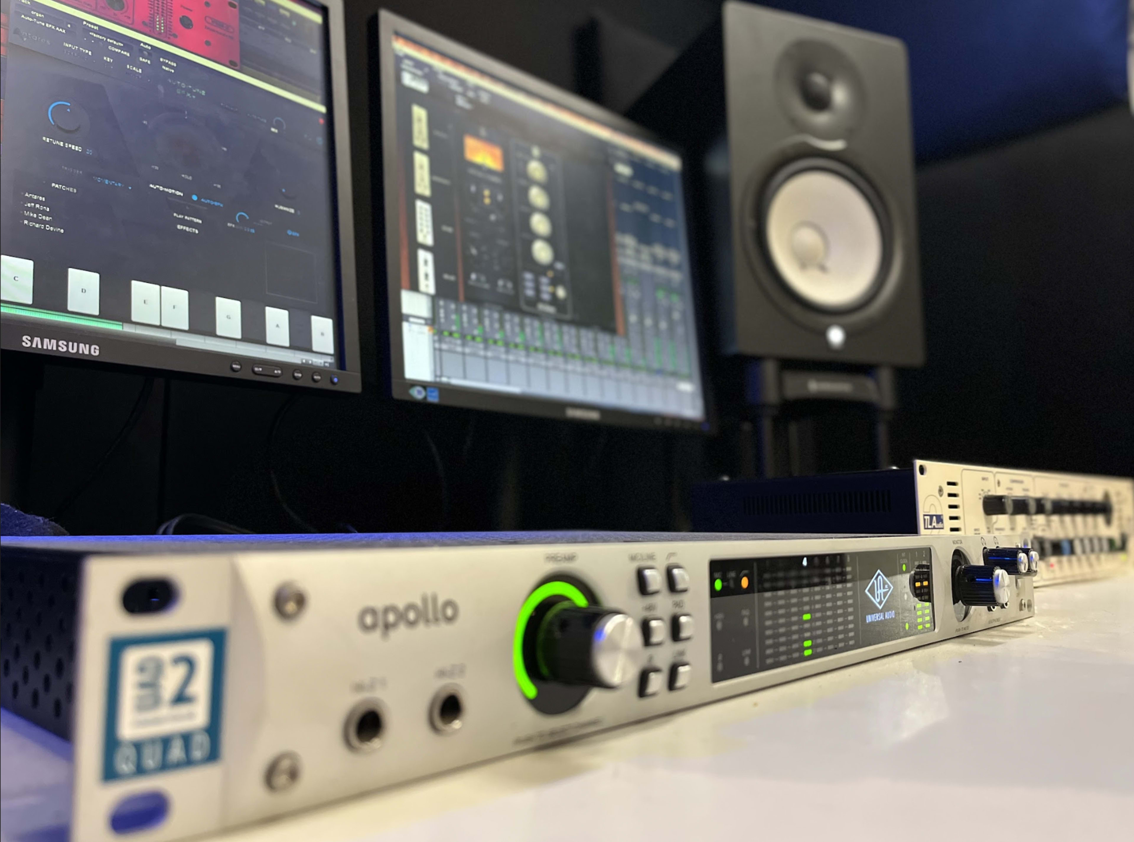 The image shows an Apollo audio interface positioned in front of two computer monitors displaying digital audio workstation software, with a studio monitor speaker in the background.