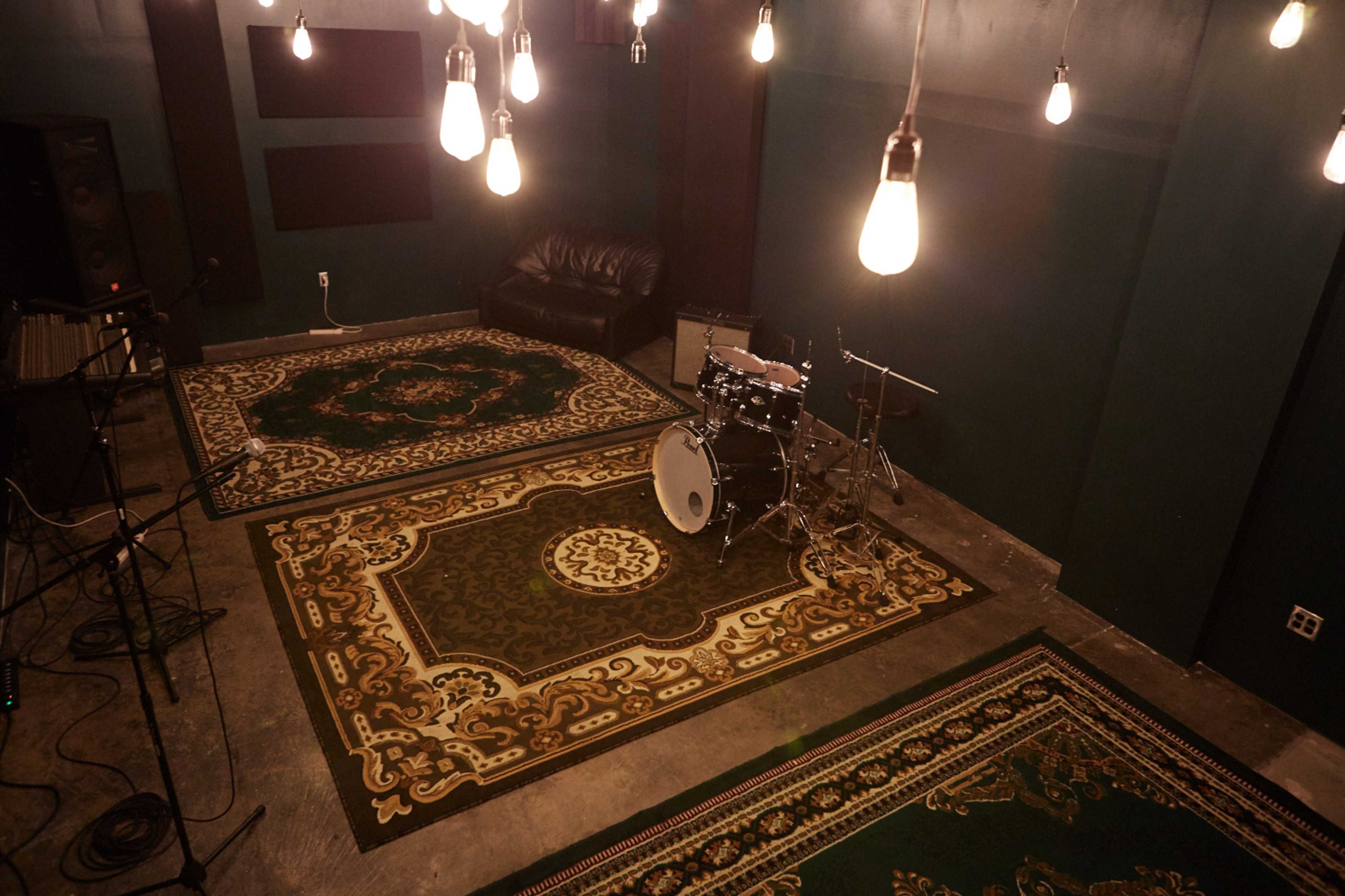 A music studio with a black couch, a drum set, and several hanging light bulbs above patterned rugs on the floor.