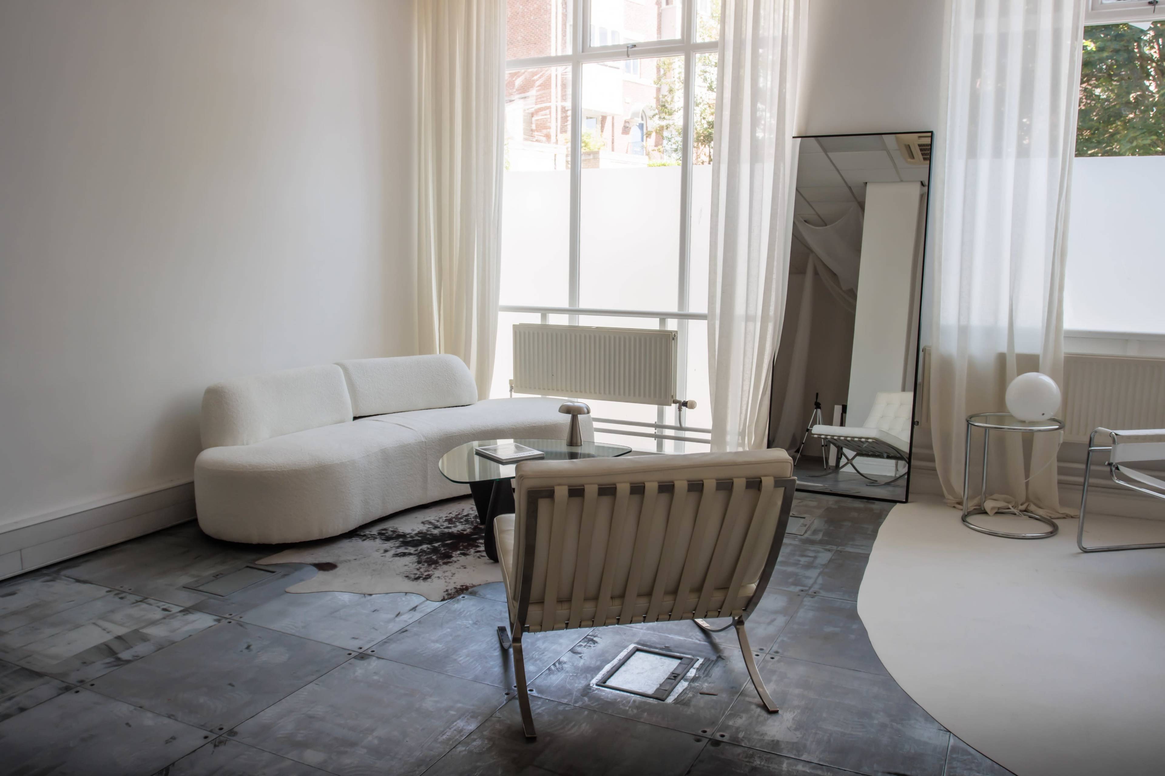 A minimalist living room featuring a curved white sofa, a glass coffee table, and a mirror, with large windows allowing natural light to fill the space.