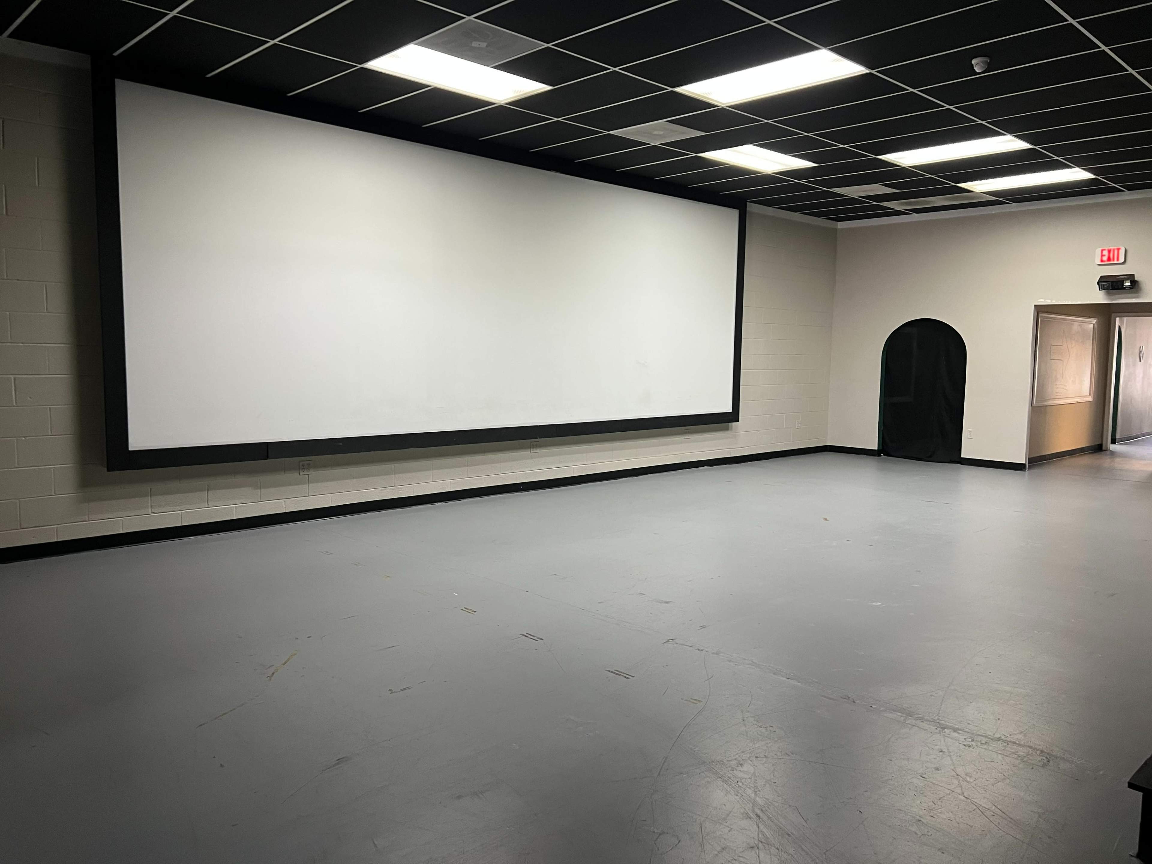 A large, blank projection screen on one wall of a sparsely furnished room with gray flooring and a black entryway on the opposite wall.