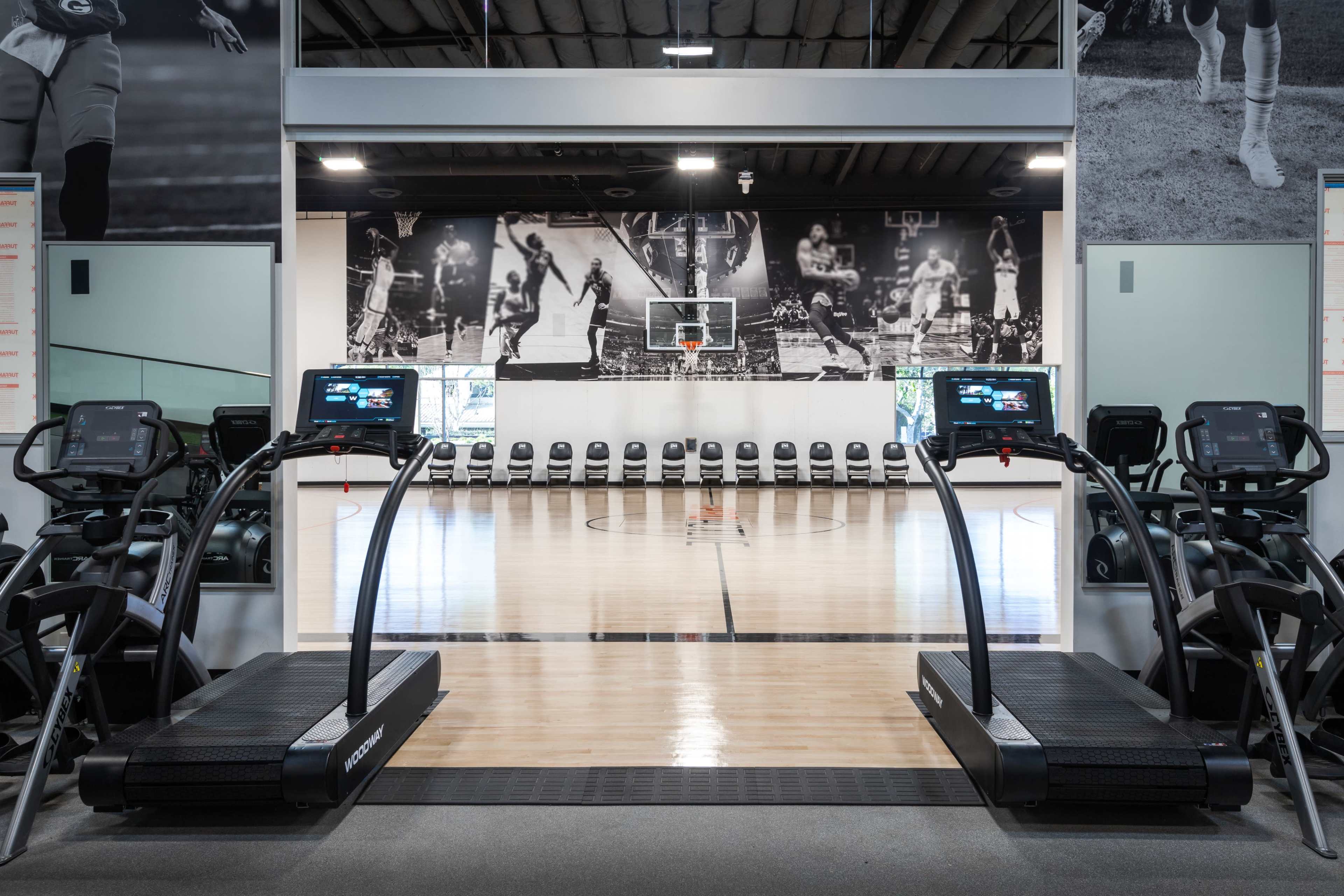 The image shows a gym with two treadmills positioned in front of a basketball court featuring a hoop and bleacher seating in the background.