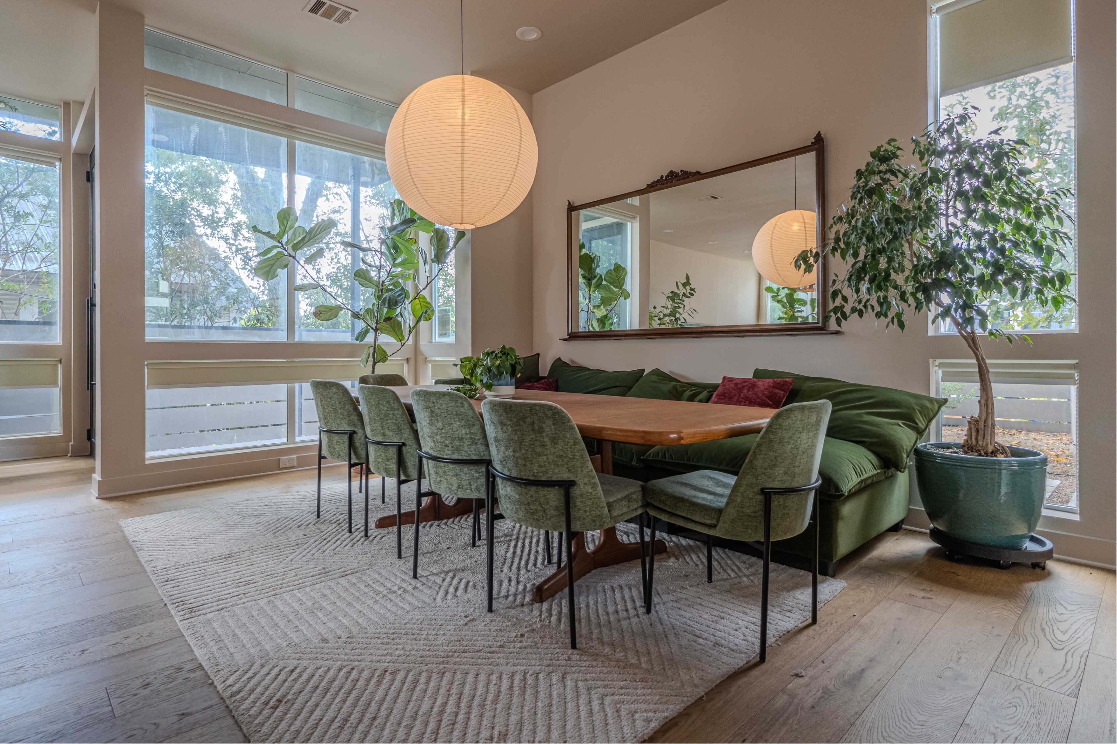 A large dining table surrounded by green chairs is positioned in a brightly lit room with large windows and indoor plants.