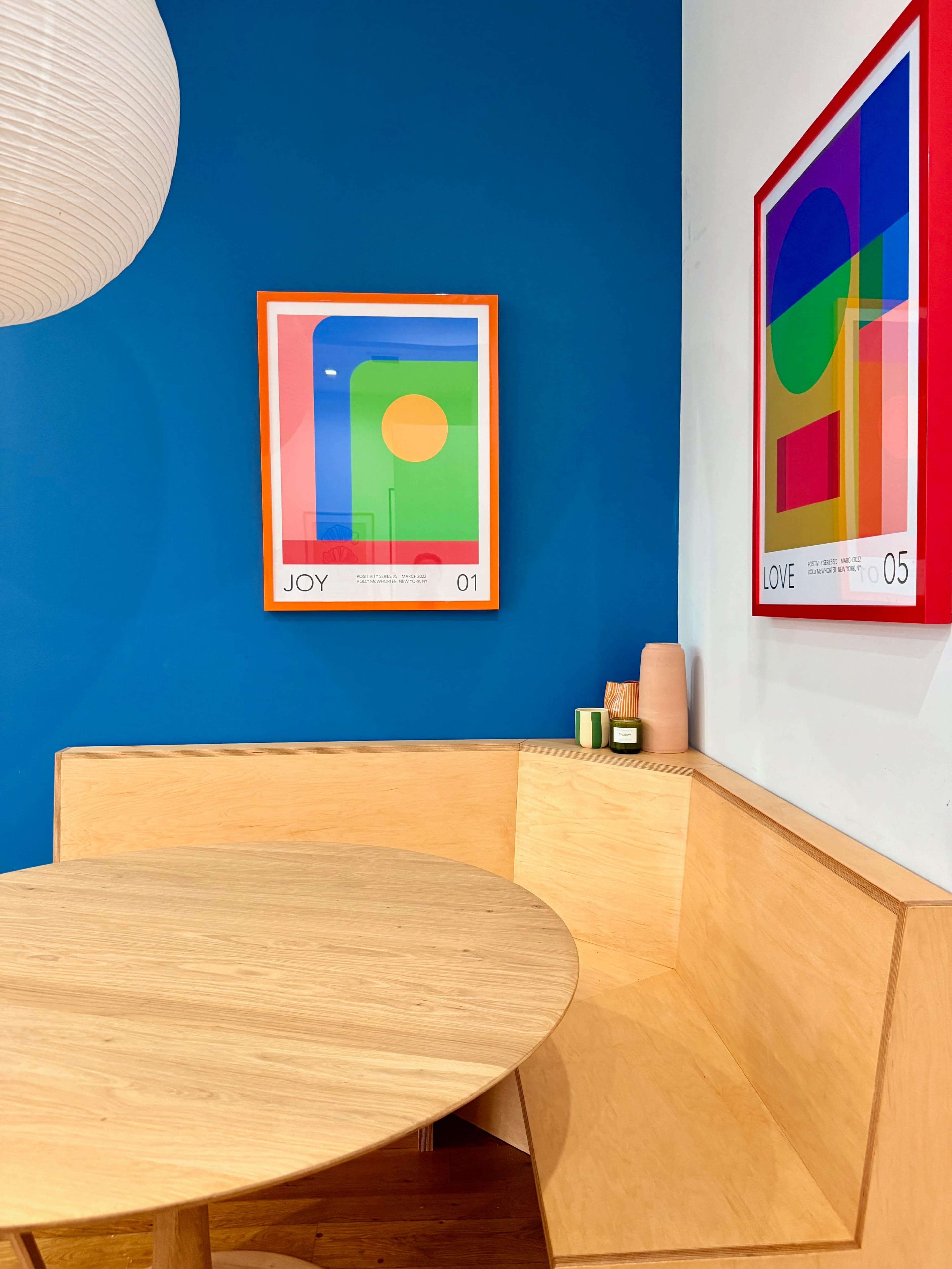 The image shows a modern corner seating area with a round wooden table, colorful artwork on the wall, and decorative items on a shelf.