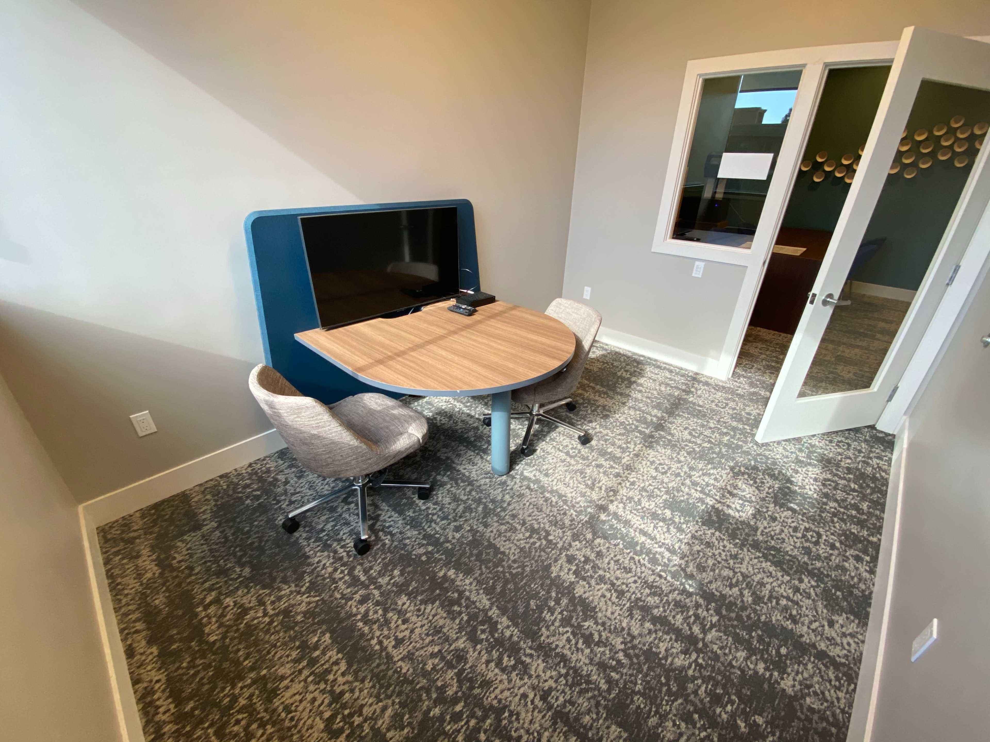 A small meeting room equipped with a round table, two swivel chairs, and a wall-mounted screen.