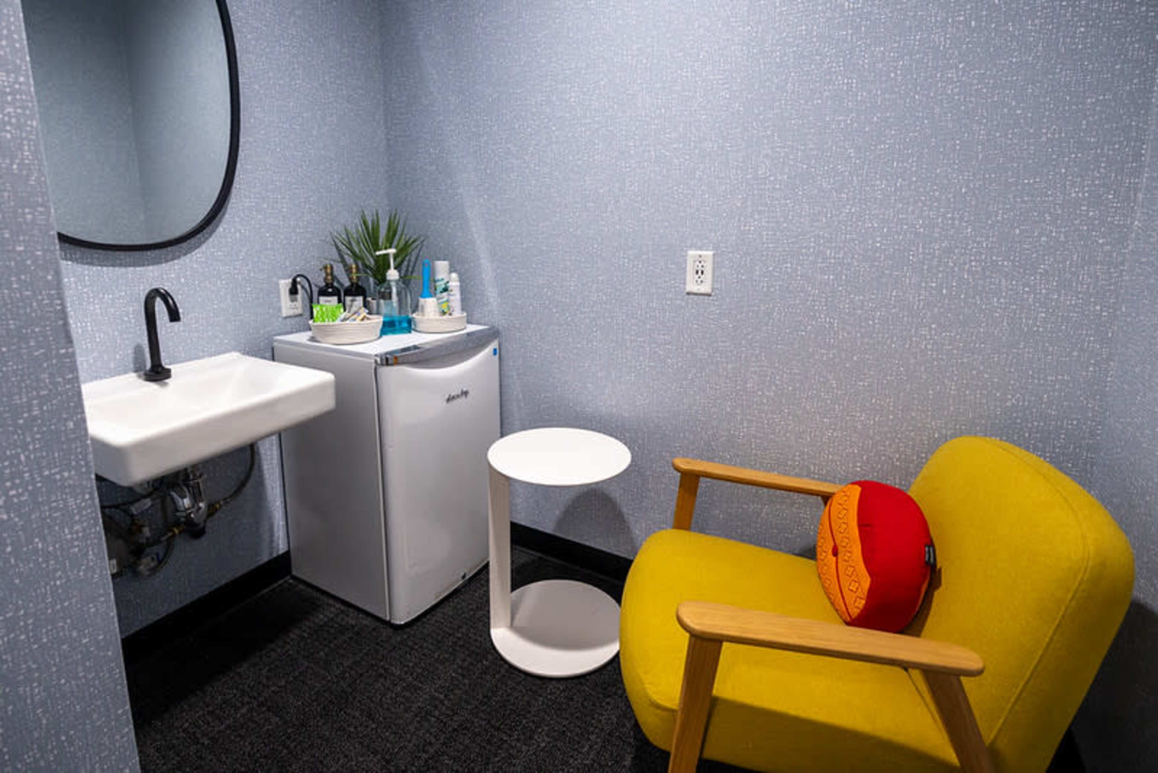 The image shows a small room featuring a yellow chair, a white side table, a mini fridge, a sink, and various toiletries on a shelf.