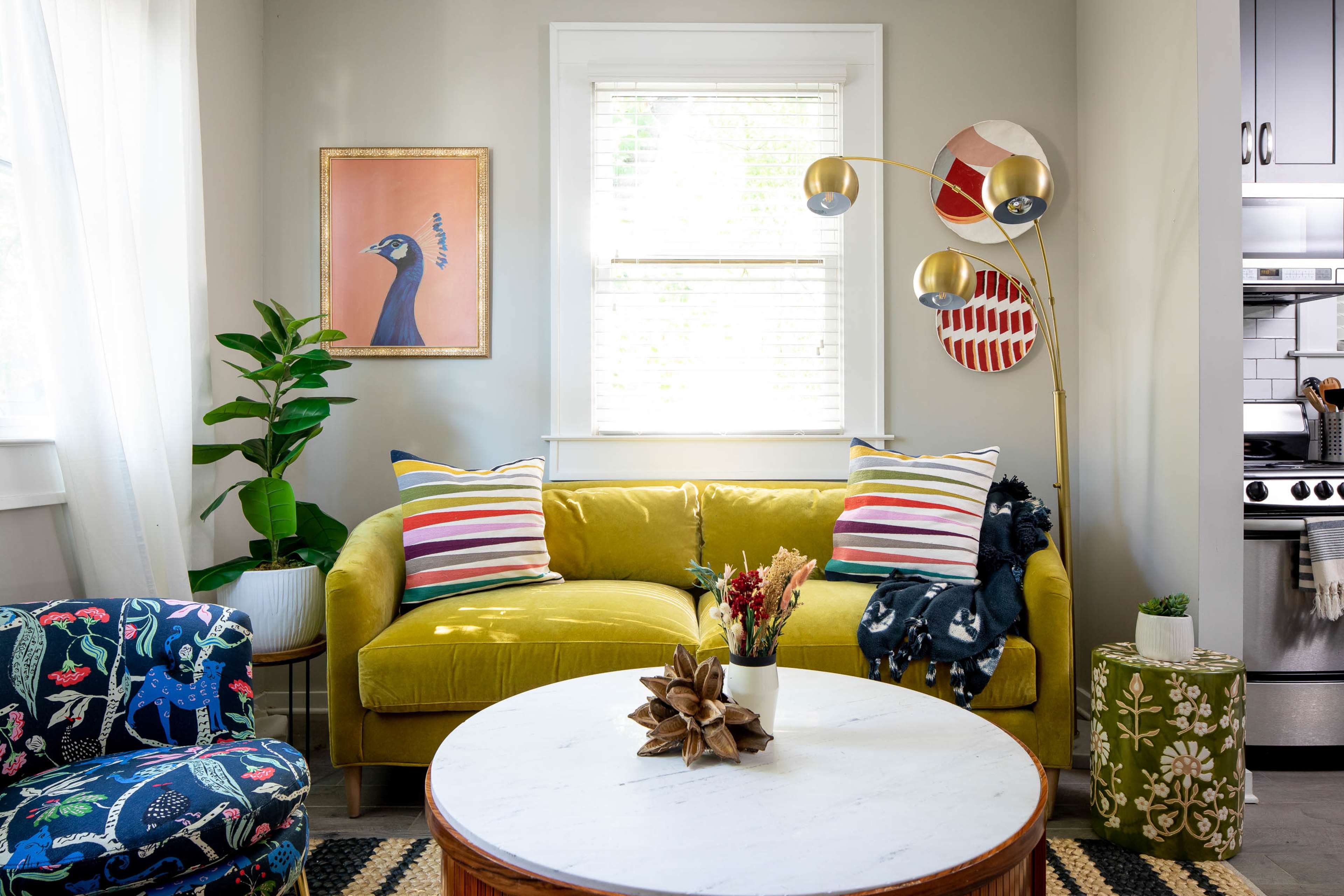 A bright living room features a yellow sofa with striped pillows, a round coffee table, a patterned armchair, and a modern floor lamp by a window.