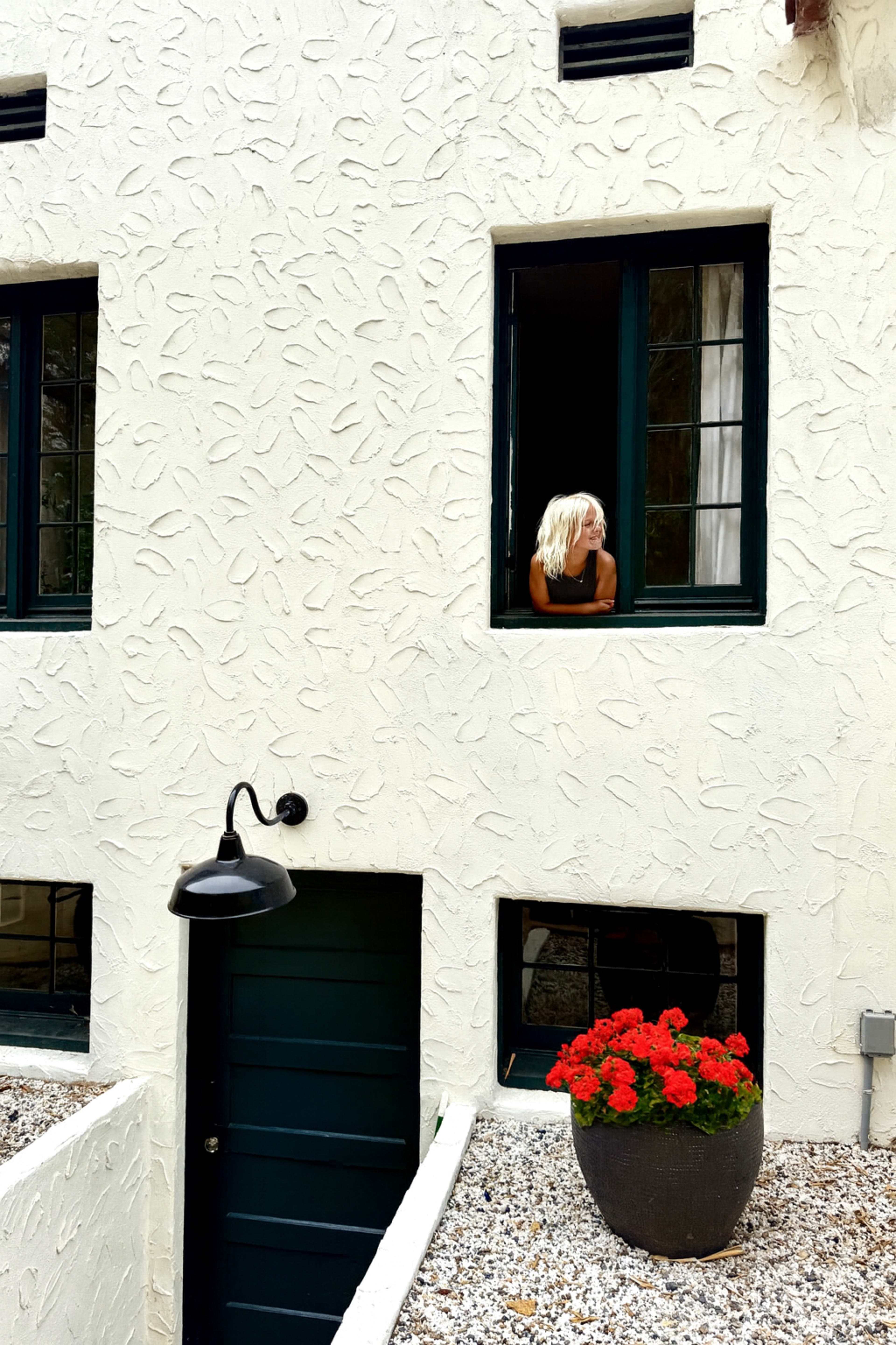 A person with long blonde hair leans out of an upper window of a white textured building, while red geraniums sit in a black planter below.