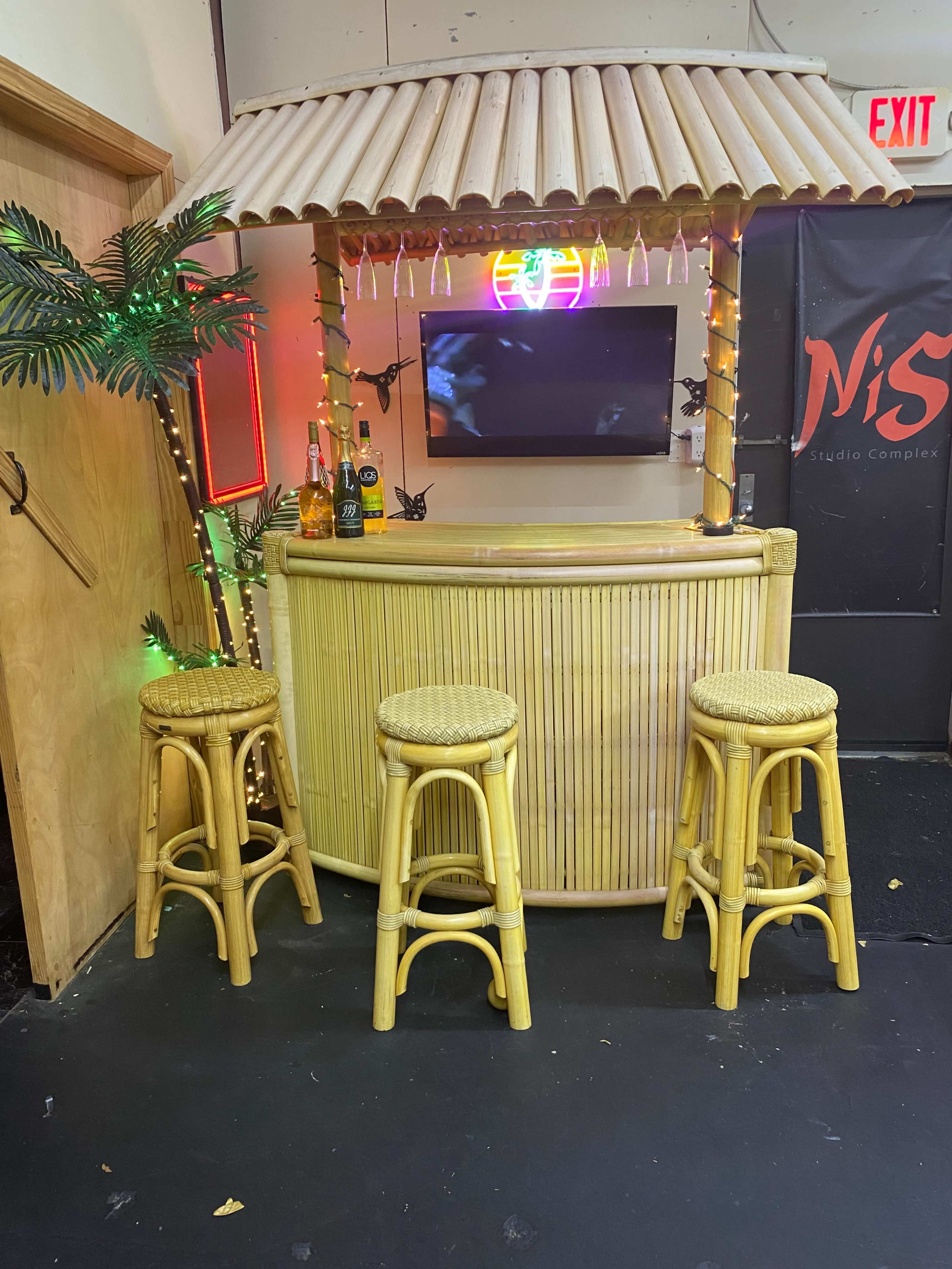 The image shows a bamboo-style bar with three straw bar stools, decorated with palm leaves and string lights, and a neon sign in the background.