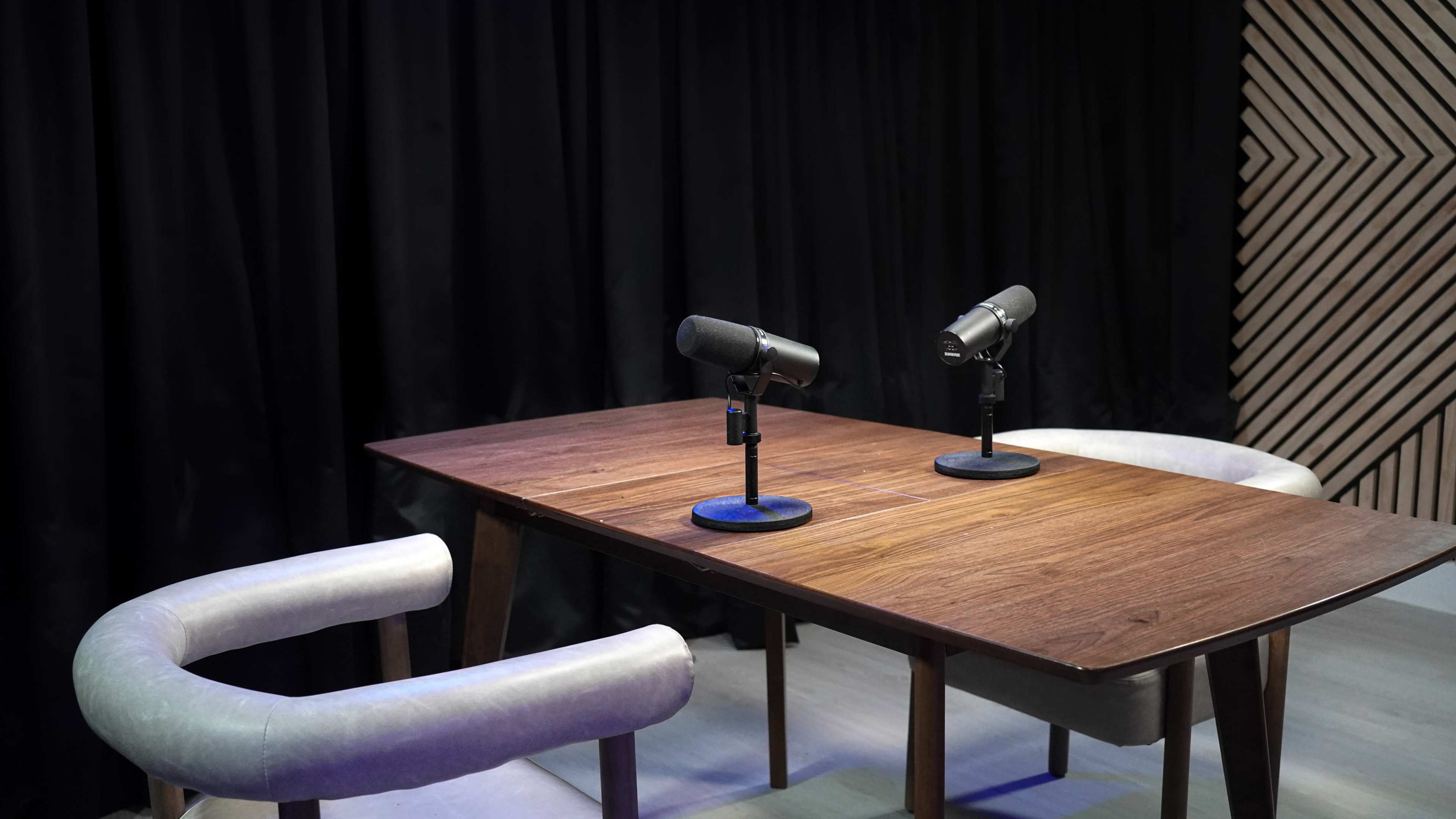 A wooden table with two microphones is set up between two chairs in a studio with a black curtain backdrop.