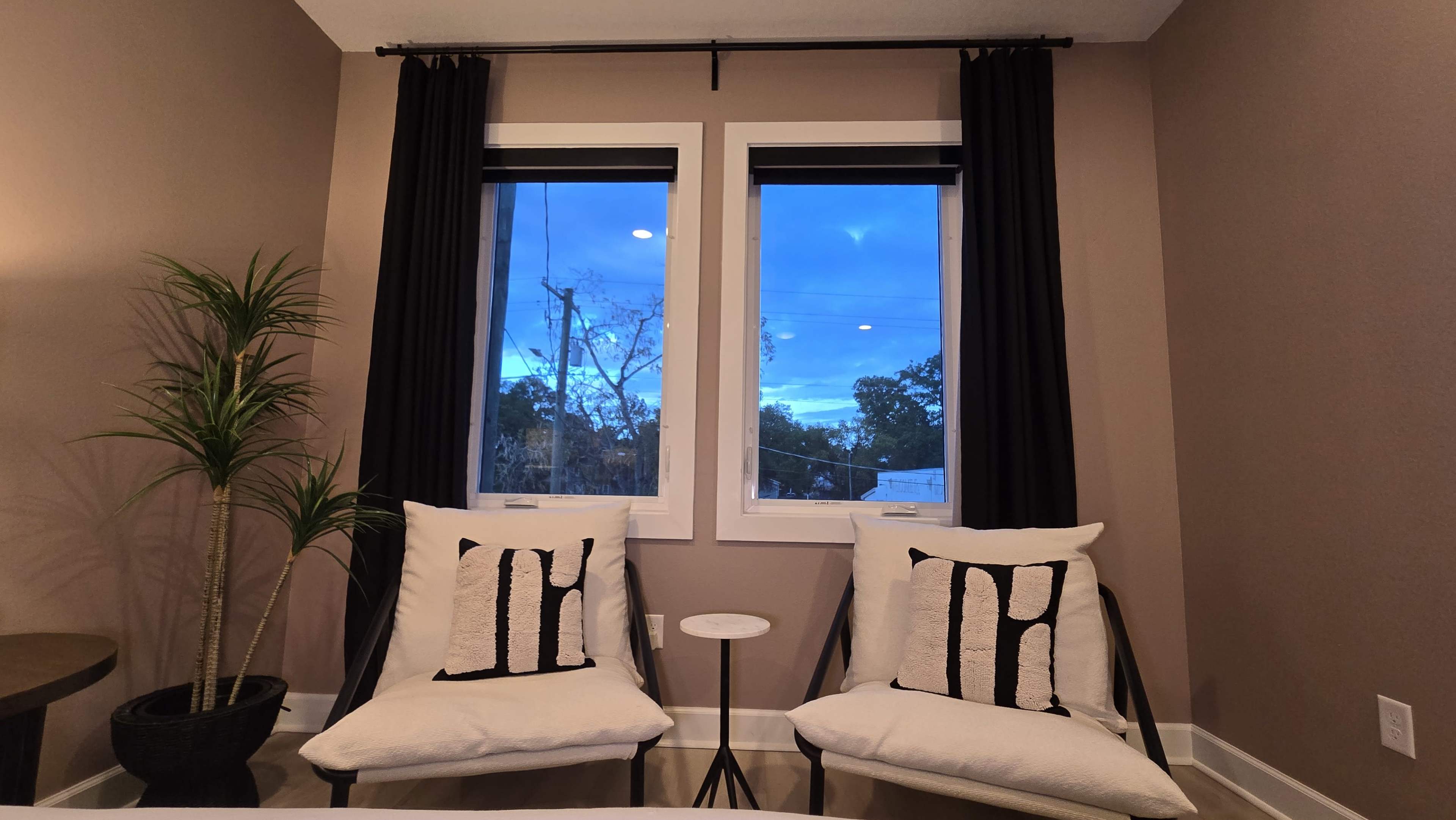 The image shows a cozy seating area with two chairs, a small table, and large windows revealing a blue sky and outdoor scenery.