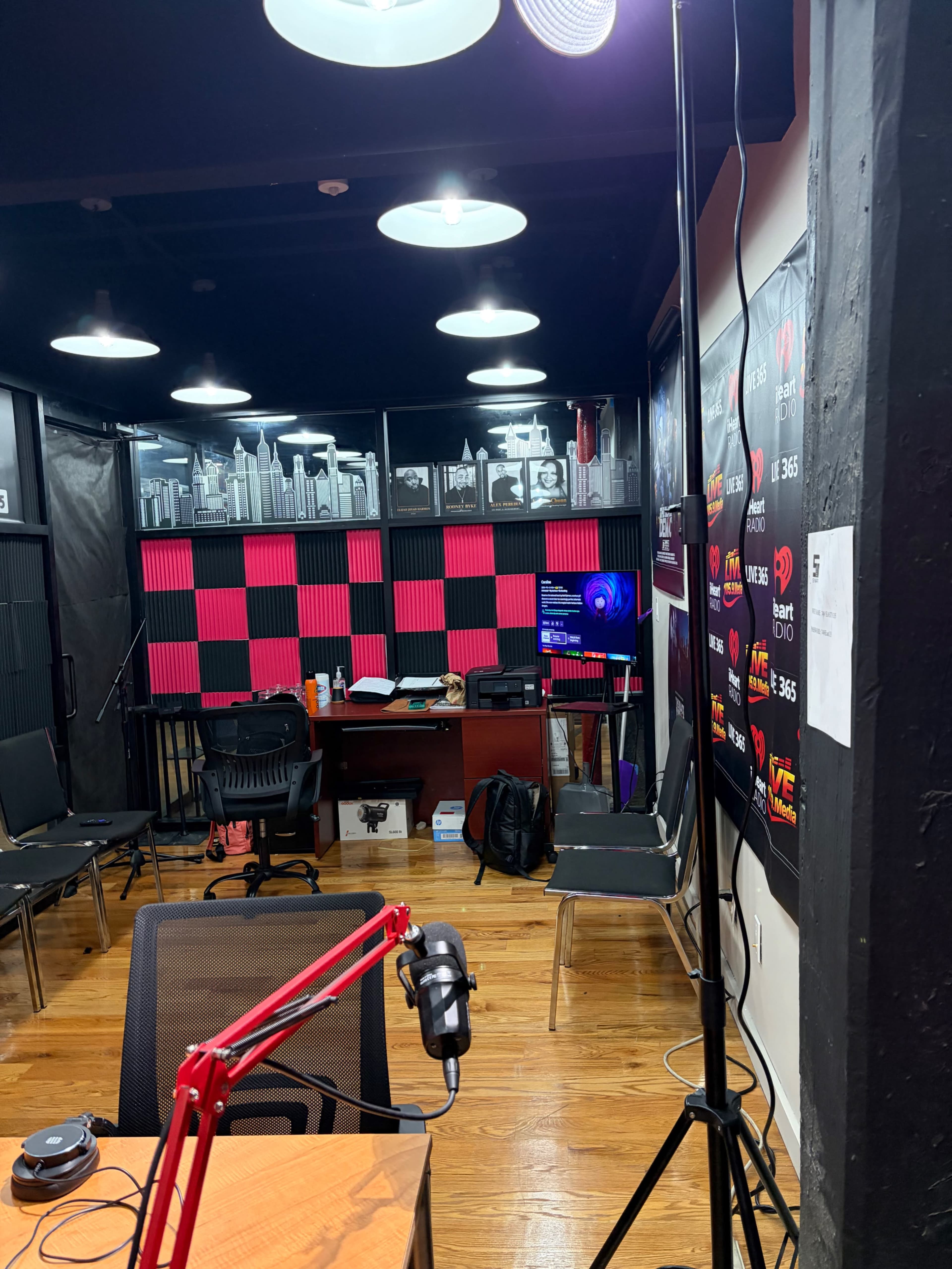 The image shows a recording studio with a microphone setup, black chairs, a desk, and walls adorned with posters and a grid pattern of pink and black.