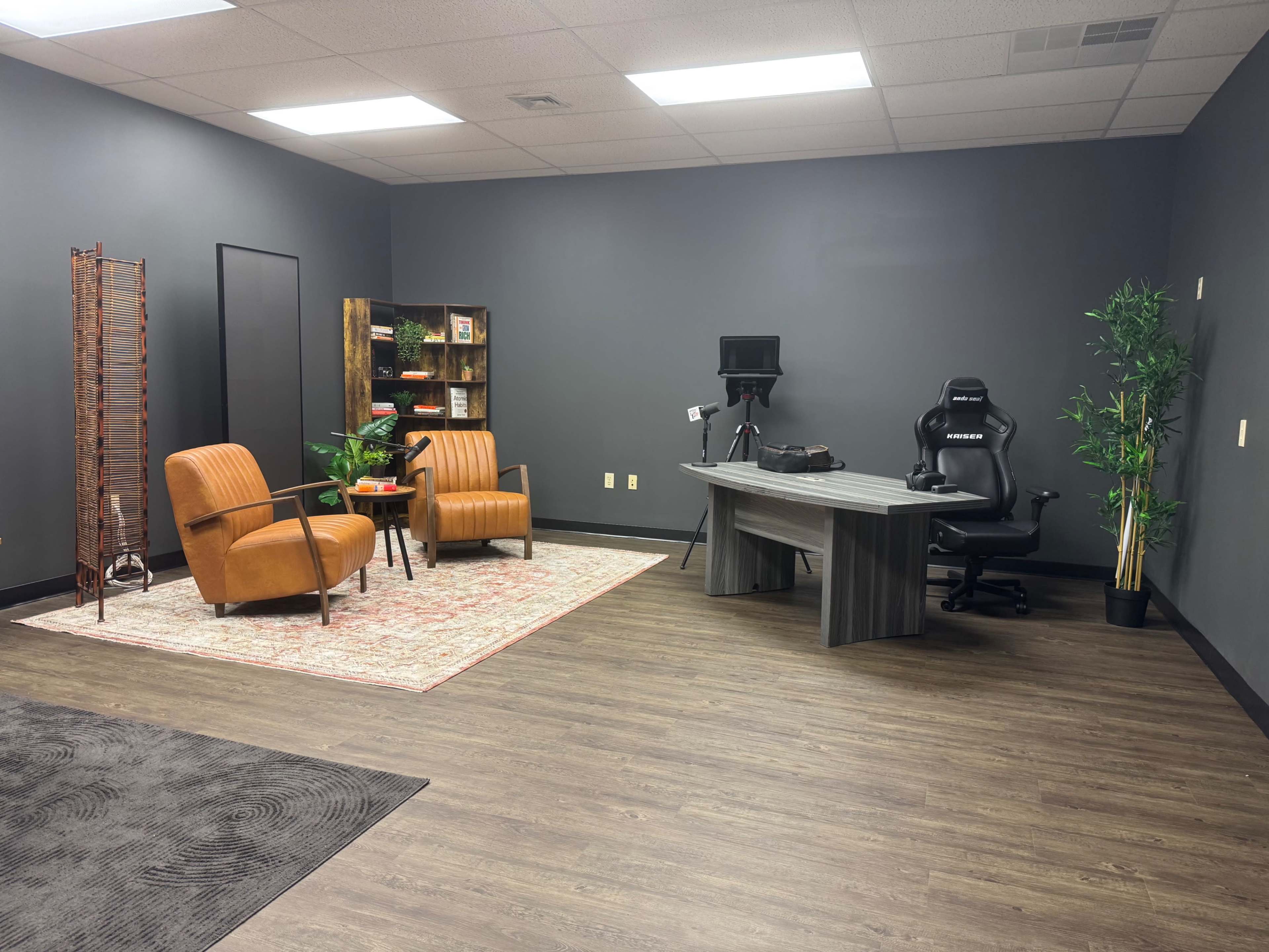 The image shows a modern office space featuring a desk with a computer chair, two orange armchairs, a bookshelf, a plant, and a backdrop for video recording.
