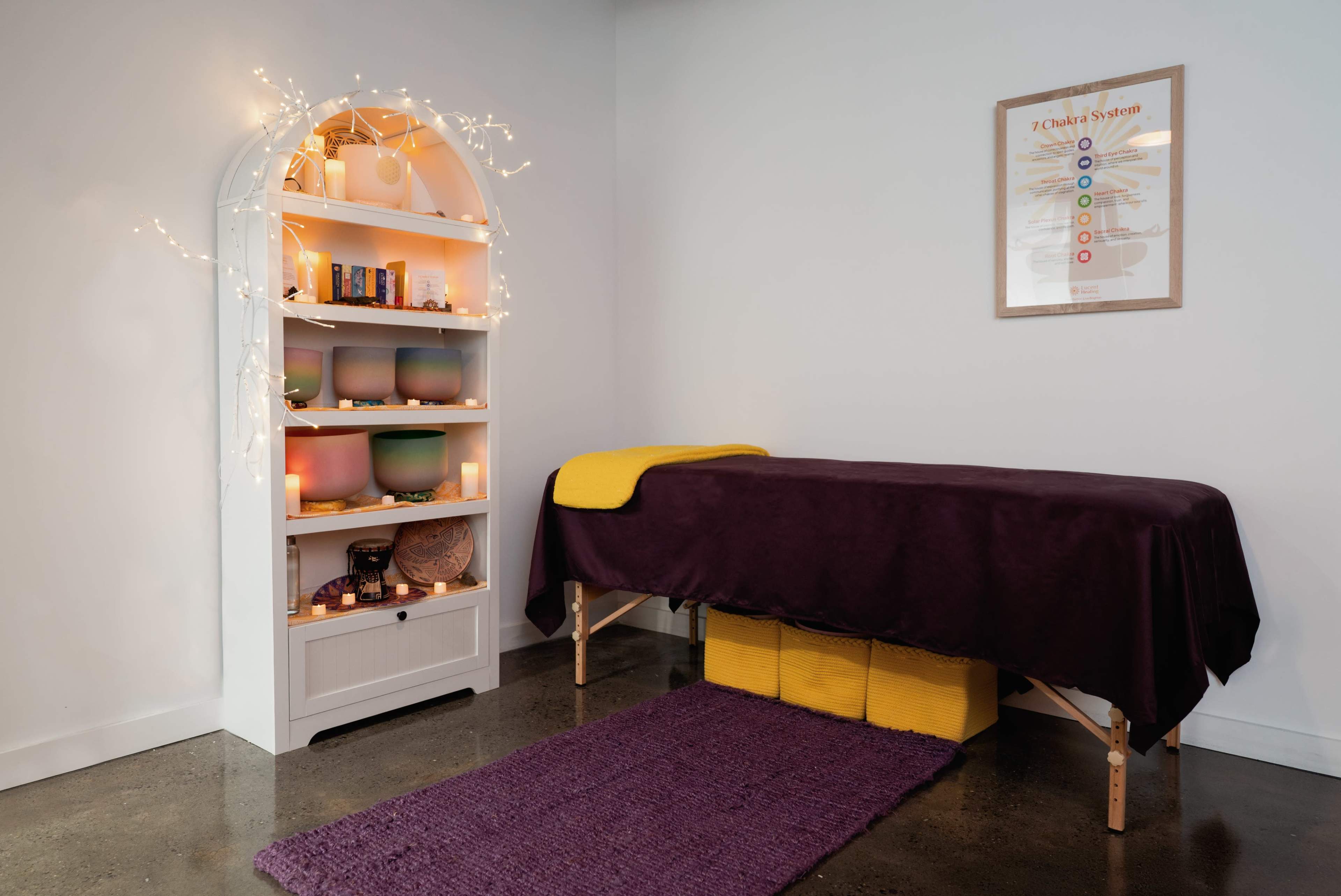 The image shows a calming wellness space with a massage table covered in a dark purple blanket, surrounded by ambient lighting and healing crystals on shelves.
