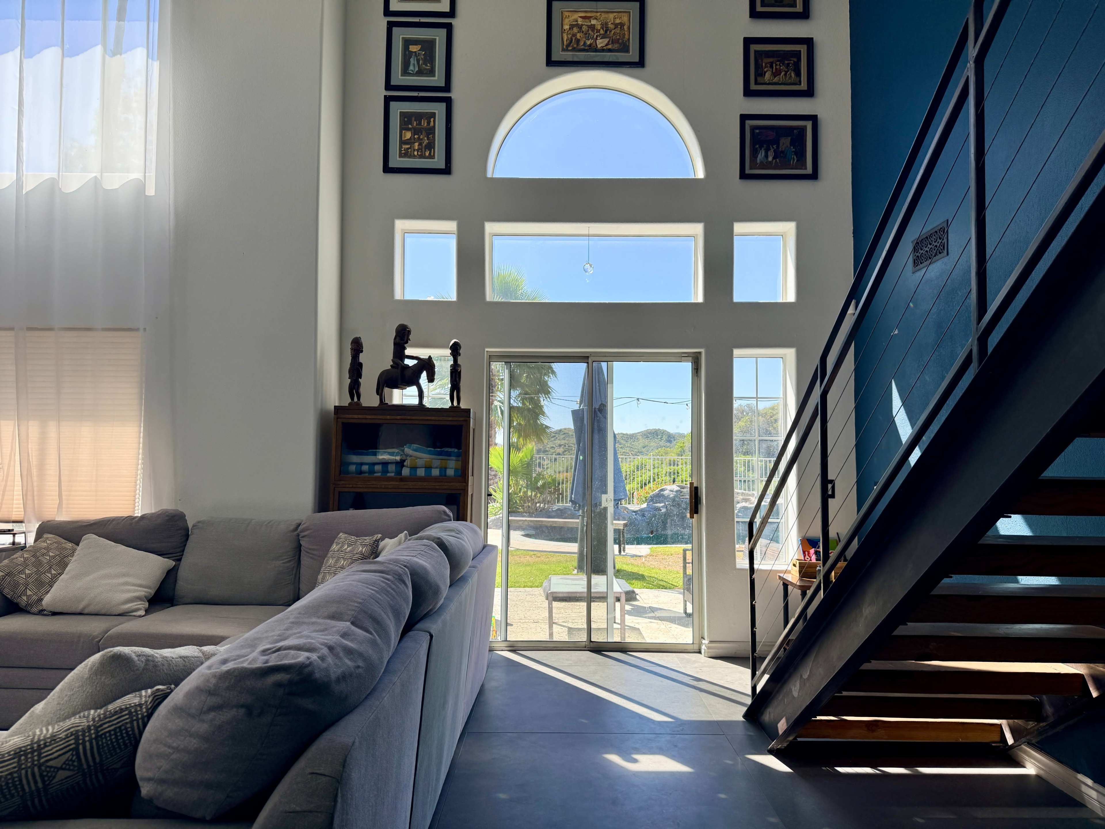 A modern living room features a large gray sofa, a staircase with metal railings, and tall windows allowing natural light to enter, revealing a view of the outdoors.