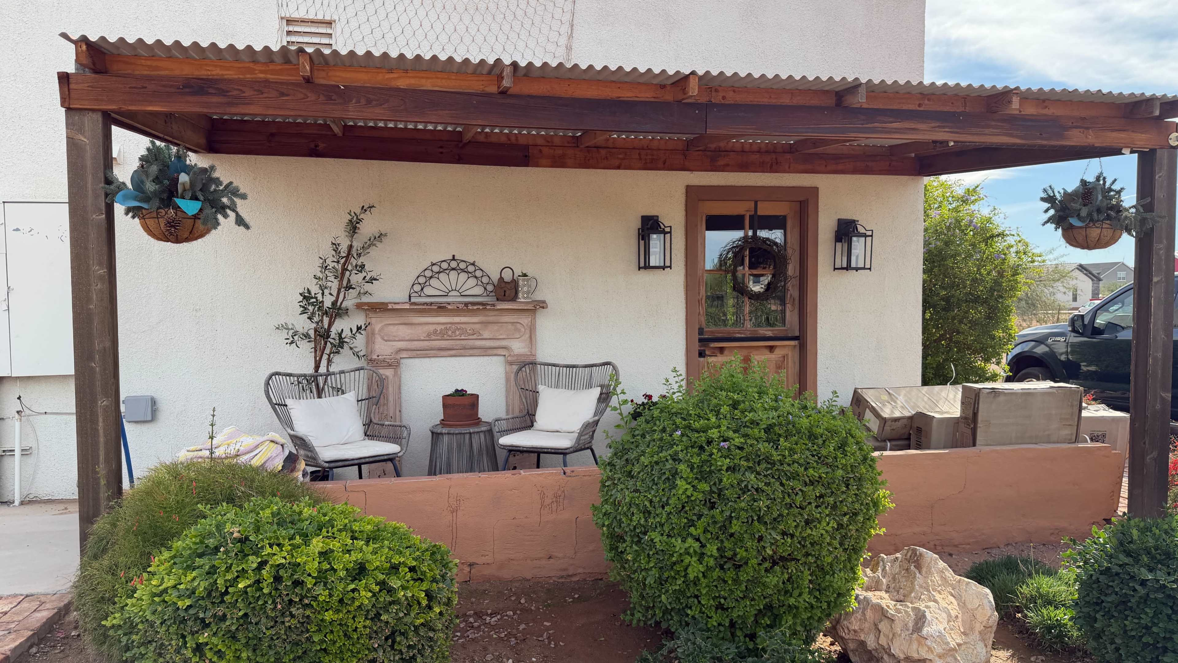The image shows a patio area with a wooden pergola, two armchairs, a small table, and a decorative stone fireplace against a white wall, surrounded by neatly trimmed bushes.