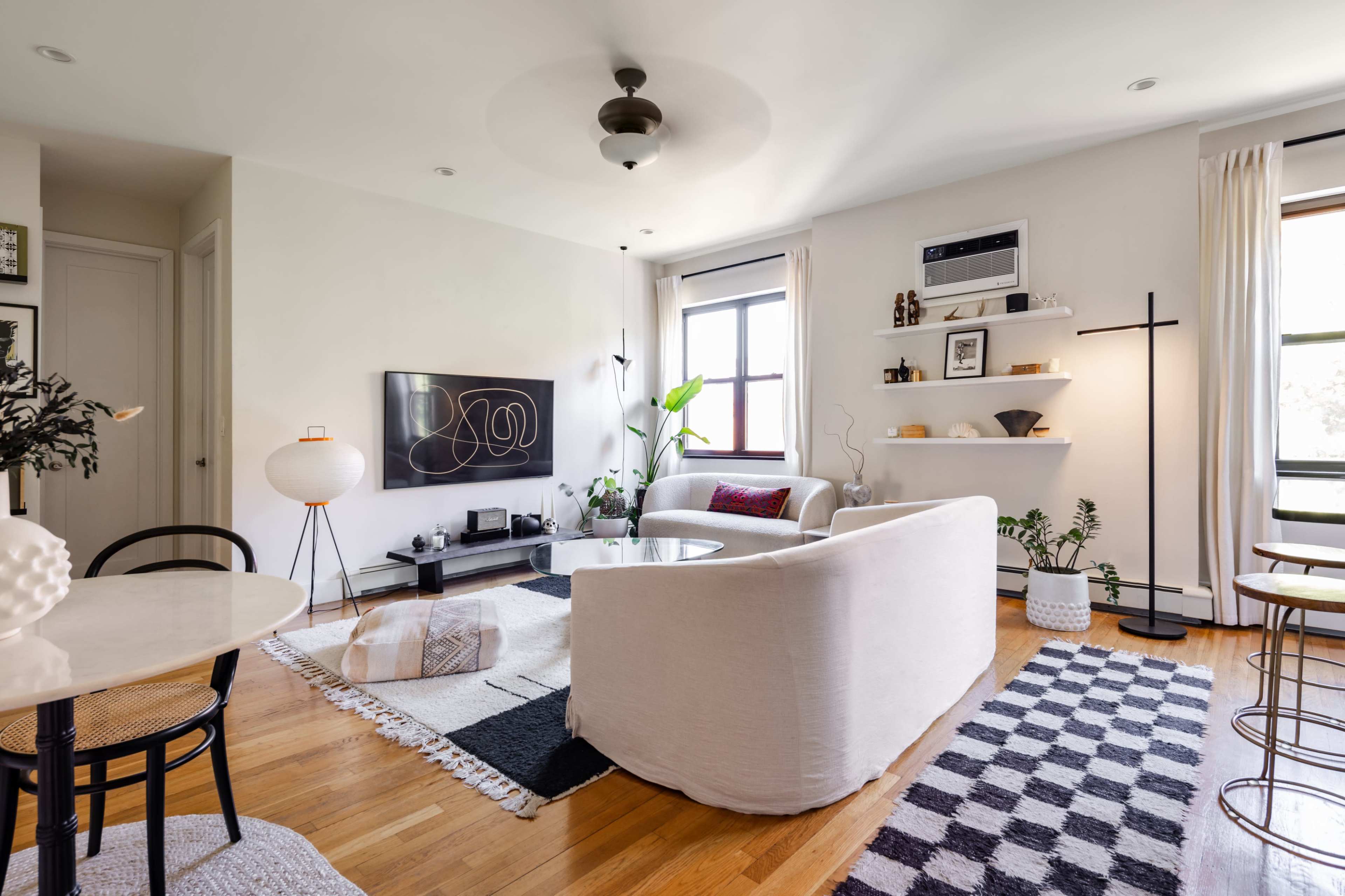 A stylish living room features a white couch, a coffee table, a television mounted on the wall, and decorative plants, with light hardwood floors and a mix of rugs.