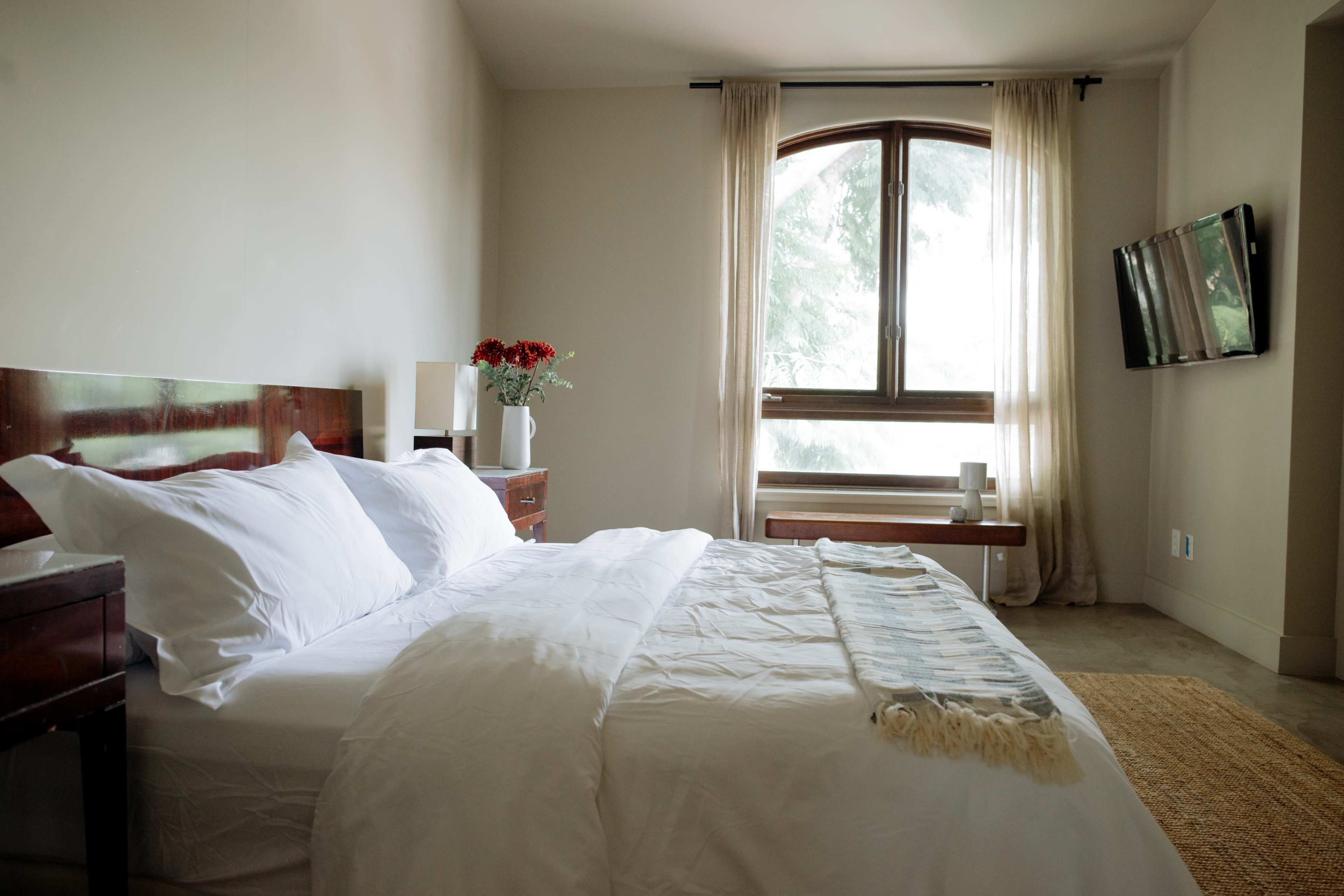 The image shows a neatly arranged bedroom with a large window, a wooden bed with white bedding, and a television mounted on the wall.