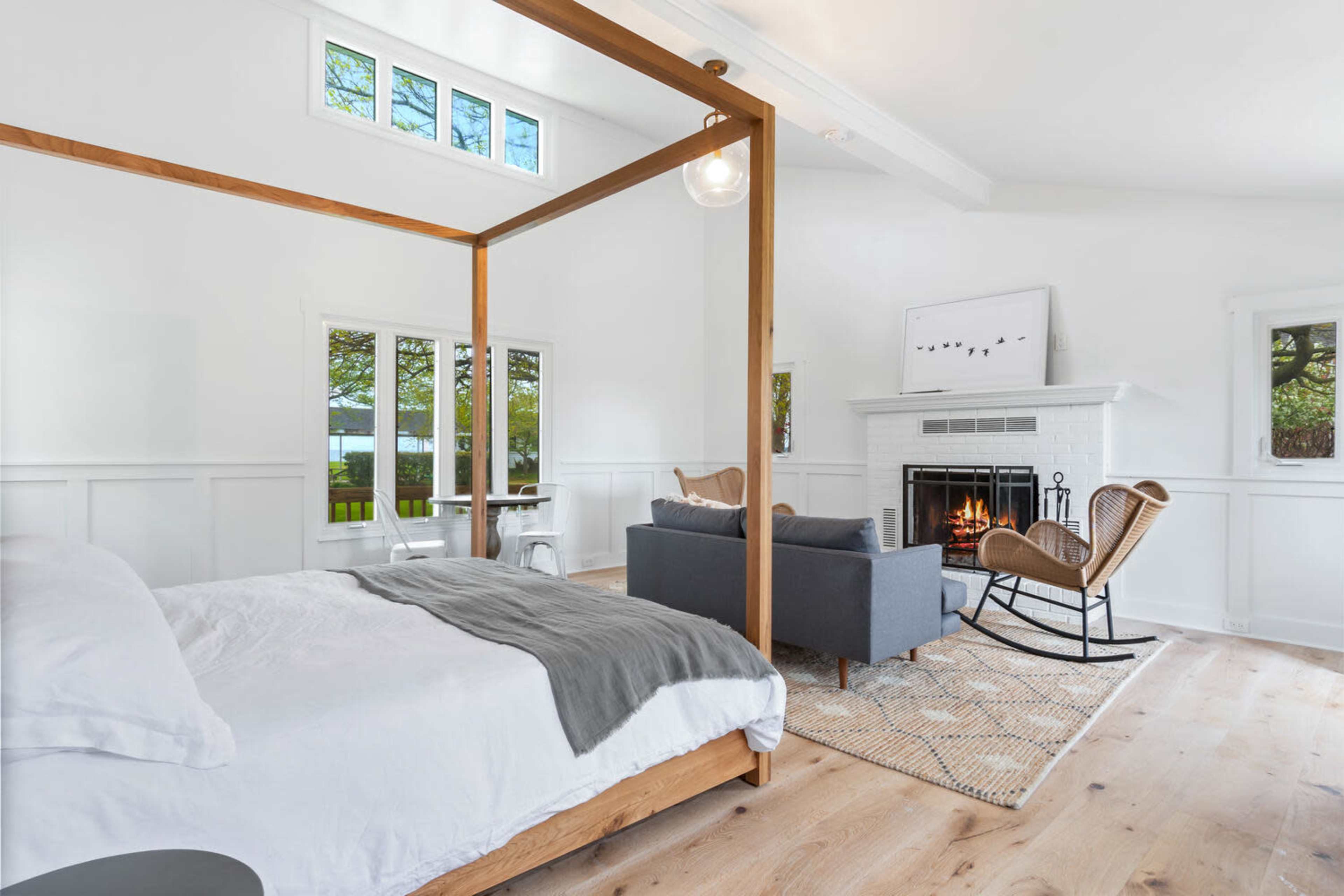 A bright, modern bedroom features a four-poster bed, a cozy seating area with a rocking chair, and a fireplace surrounded by white walls and large windows.