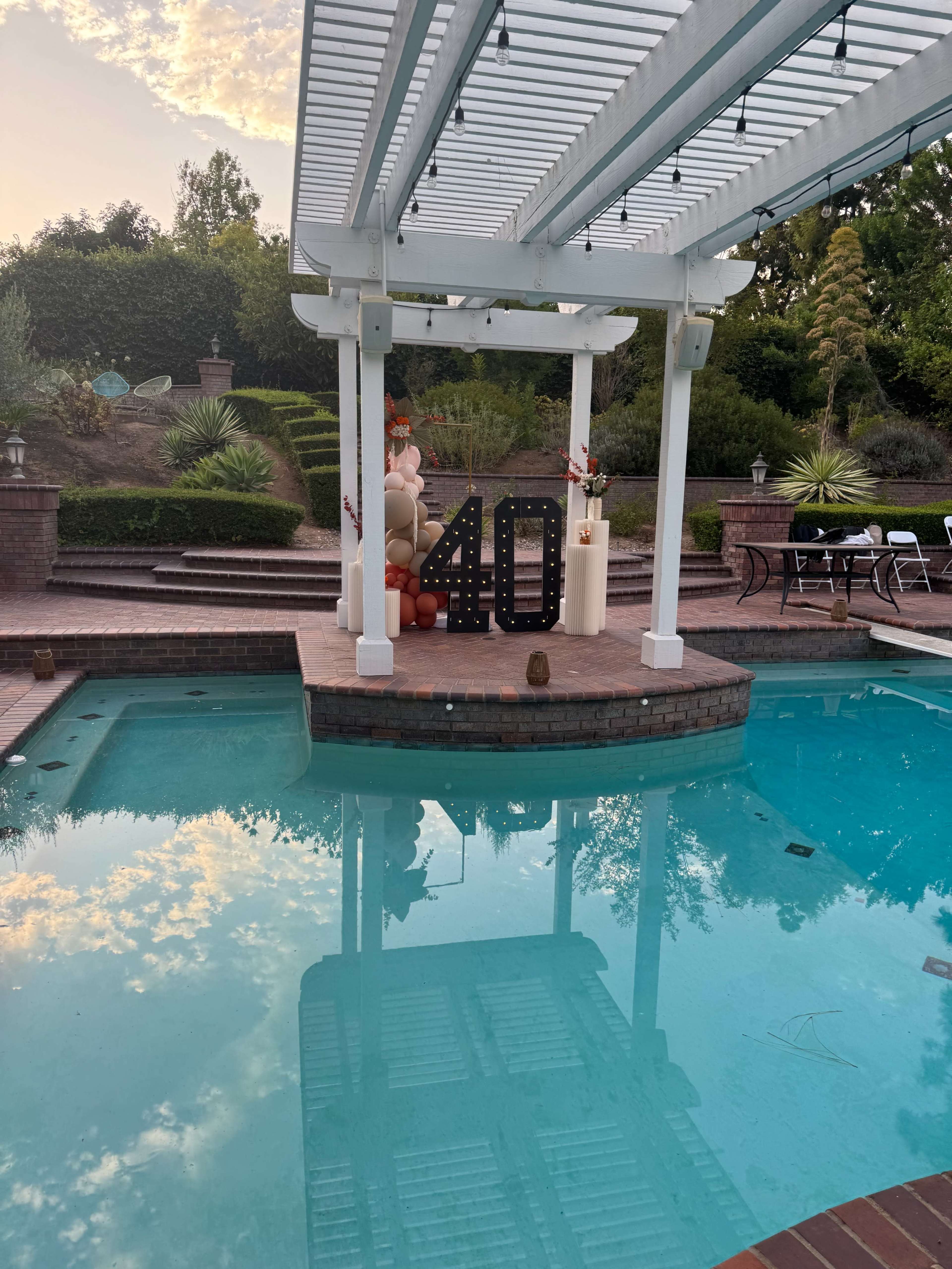 The image shows a pool area with a decorative setup featuring the number "40" and balloons under a pergola, surrounded by landscaped gardens.