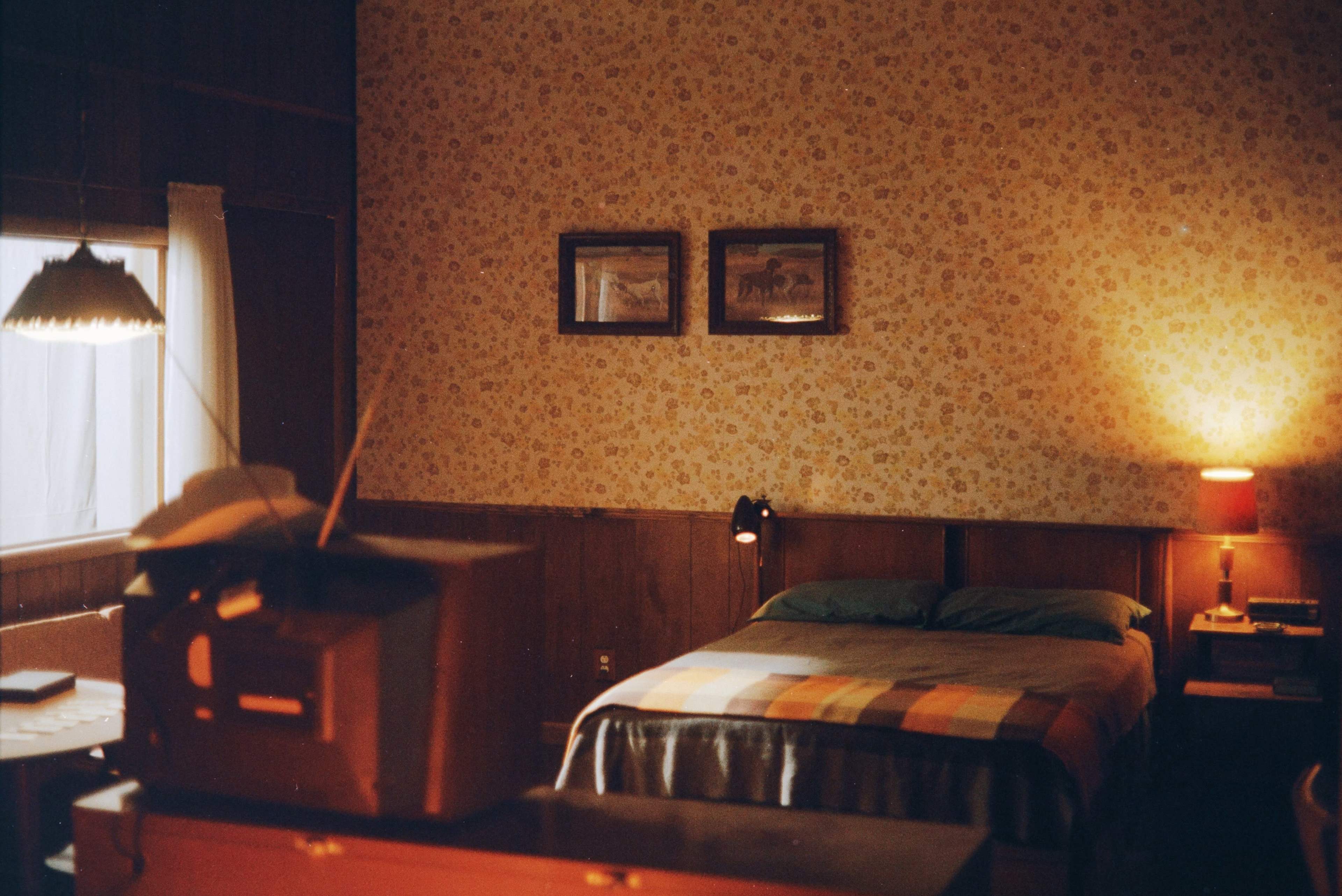 The image shows a bedroom with floral wallpaper, a bed covered with a plaid blanket, framed pictures on the wall, and a vintage television set on a dresser.