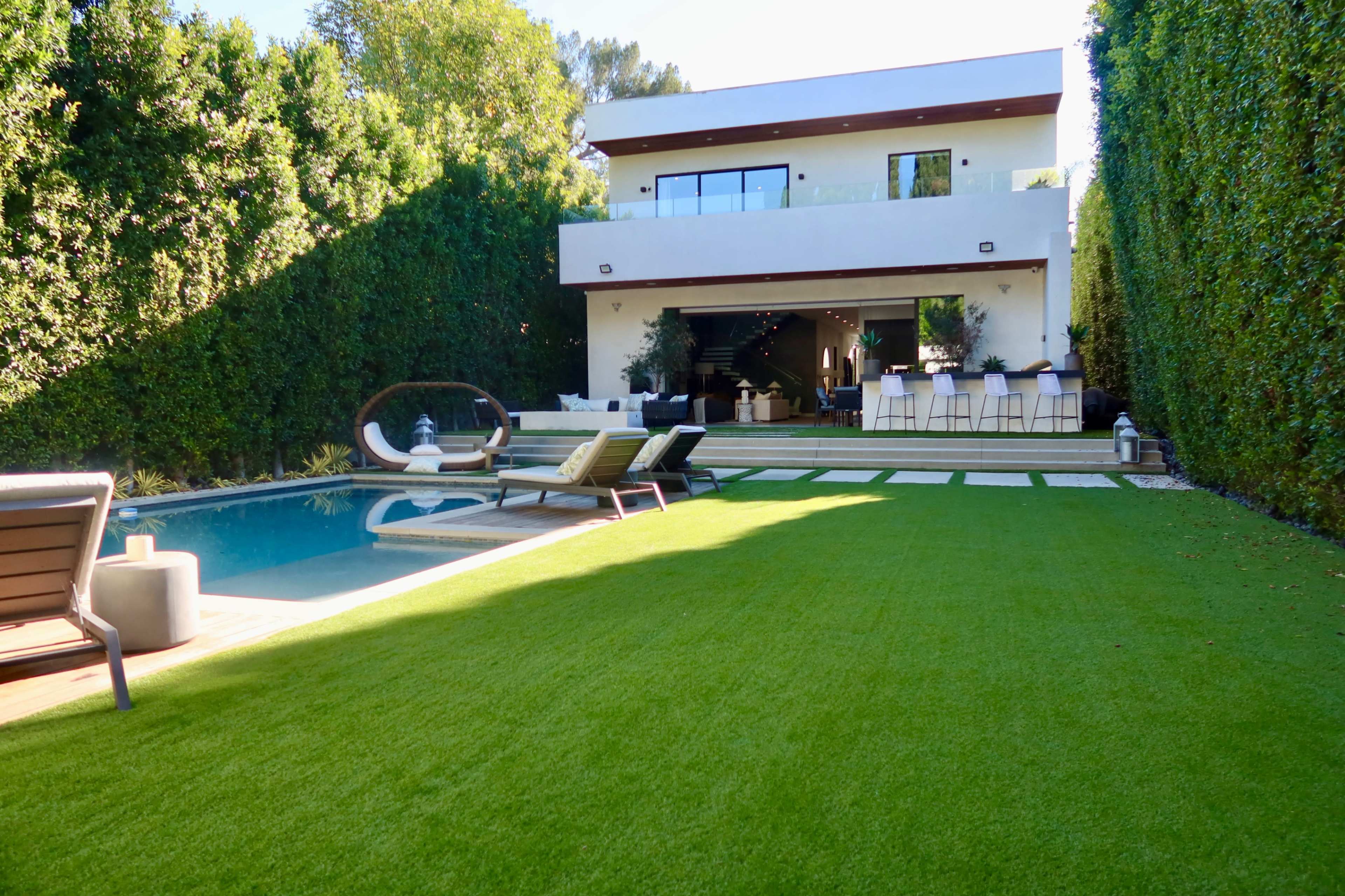 A modern two-story house with large windows overlooks a neatly landscaped backyard featuring a swimming pool and lounging area.