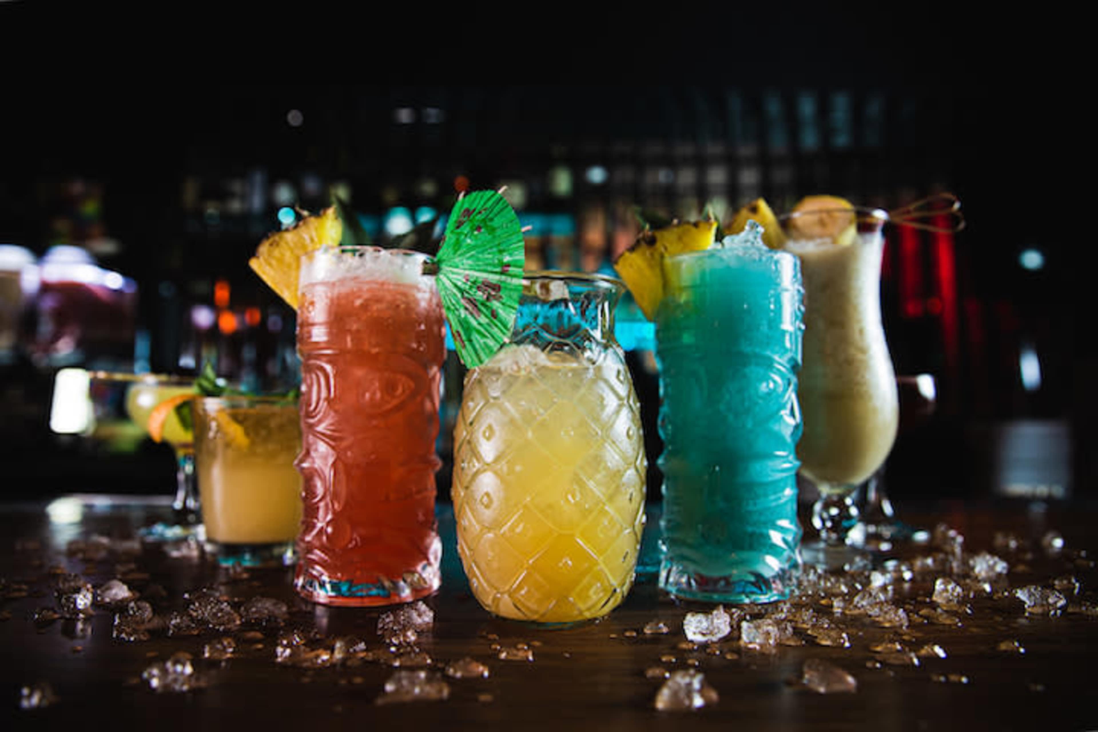 A variety of colorful cocktails are displayed on a bar counter, surrounded by scattered ice and garnished with fruit and umbrellas.