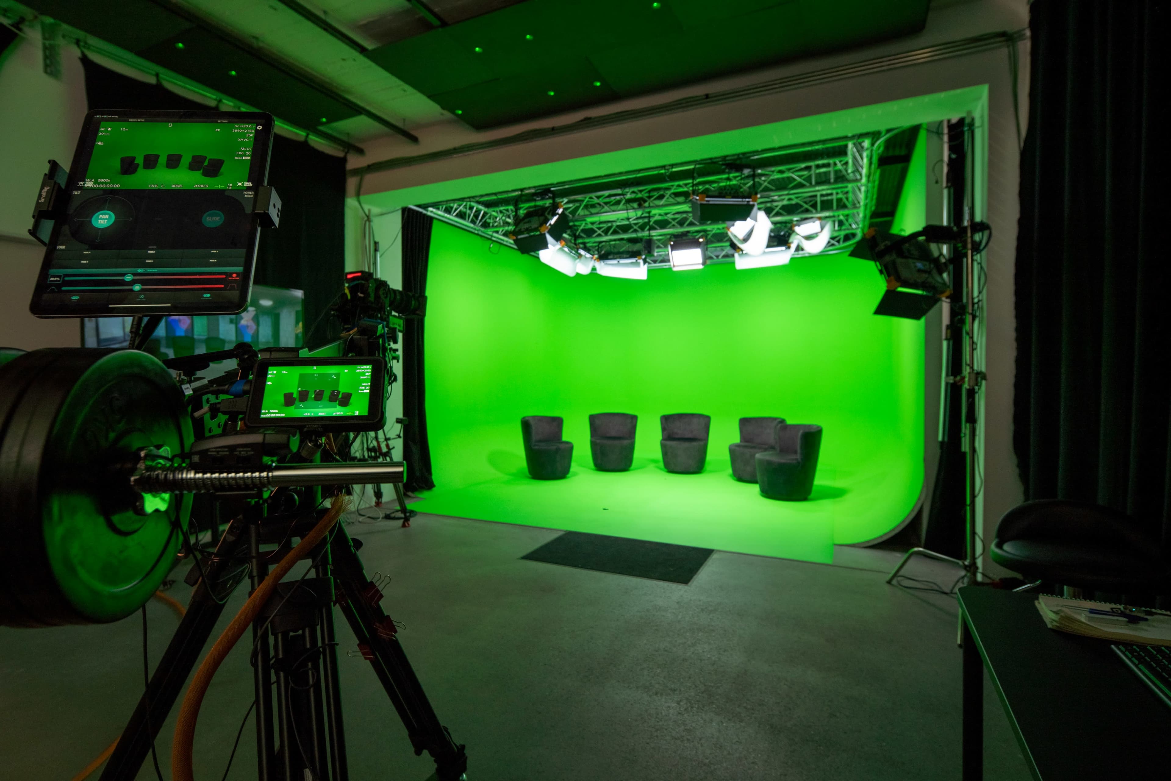 A film studio setup featuring a green screen backdrop, four black chairs, and various lighting and camera equipment.