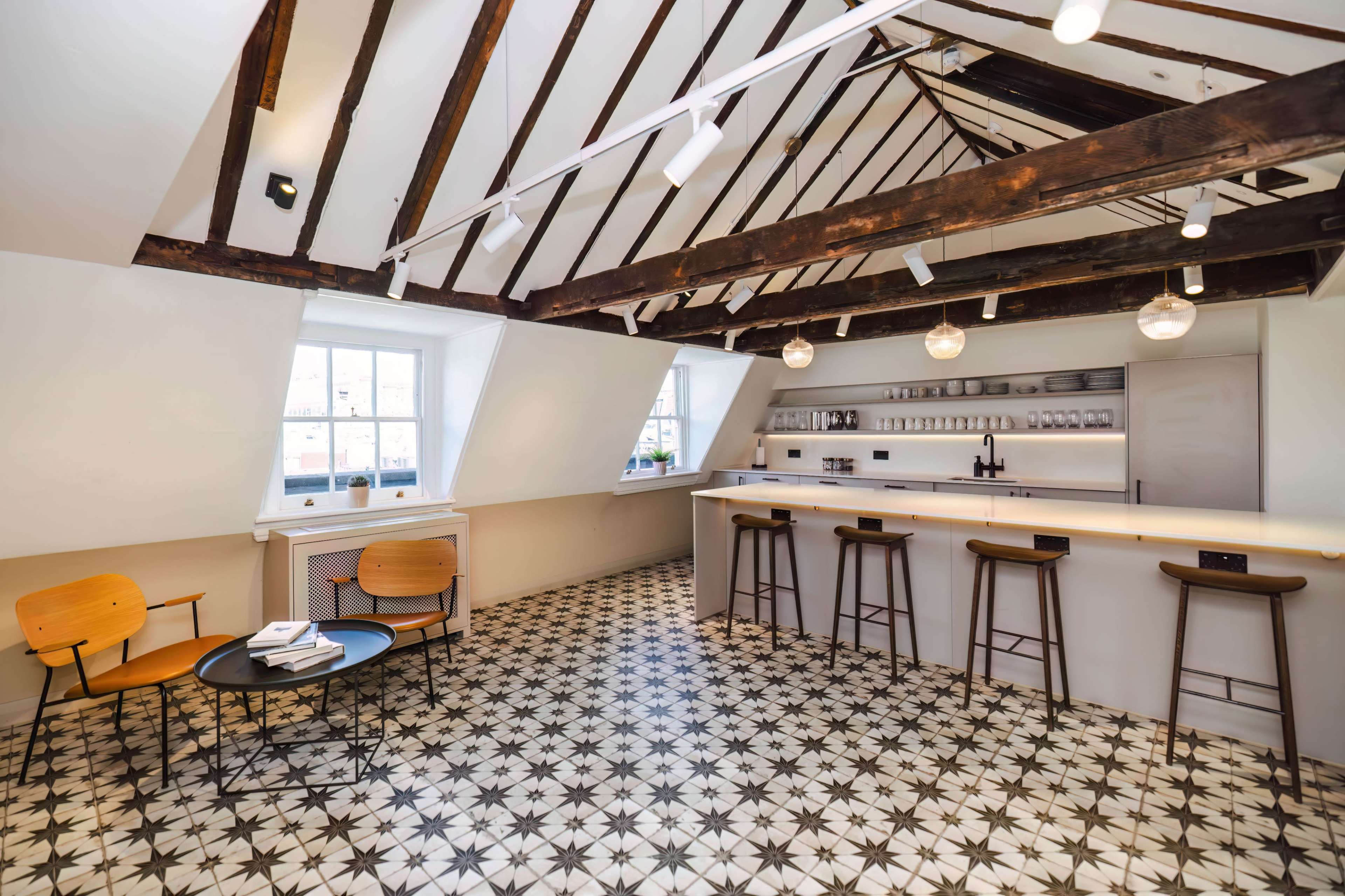 A modern café with a bar counter, wooden beams on the ceiling, and patterned tile flooring, furnished with two chairs and a small table.