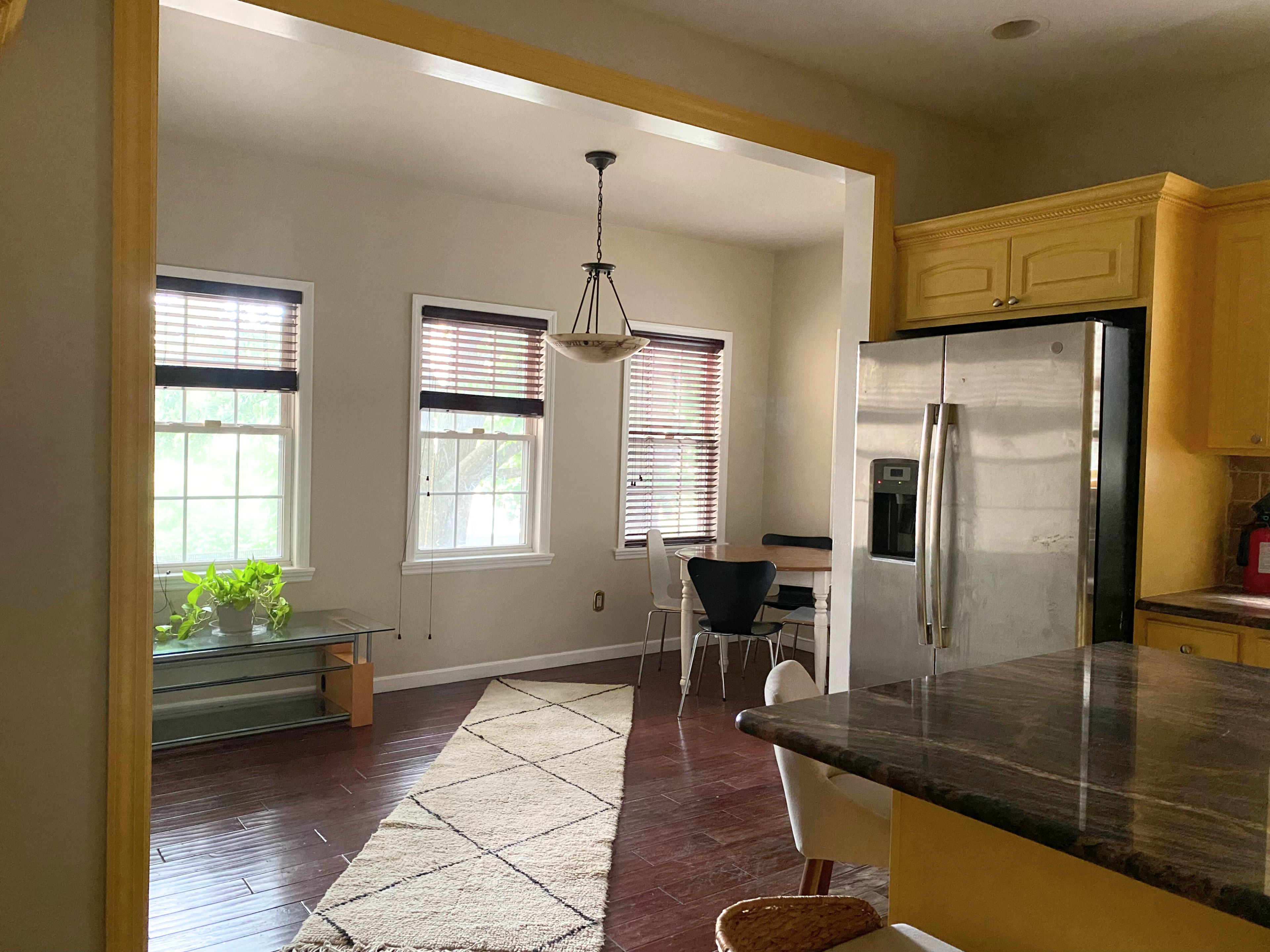 A well-lit kitchen features a dining area with a small table and chairs, surrounded by large windows and a patterned rug.