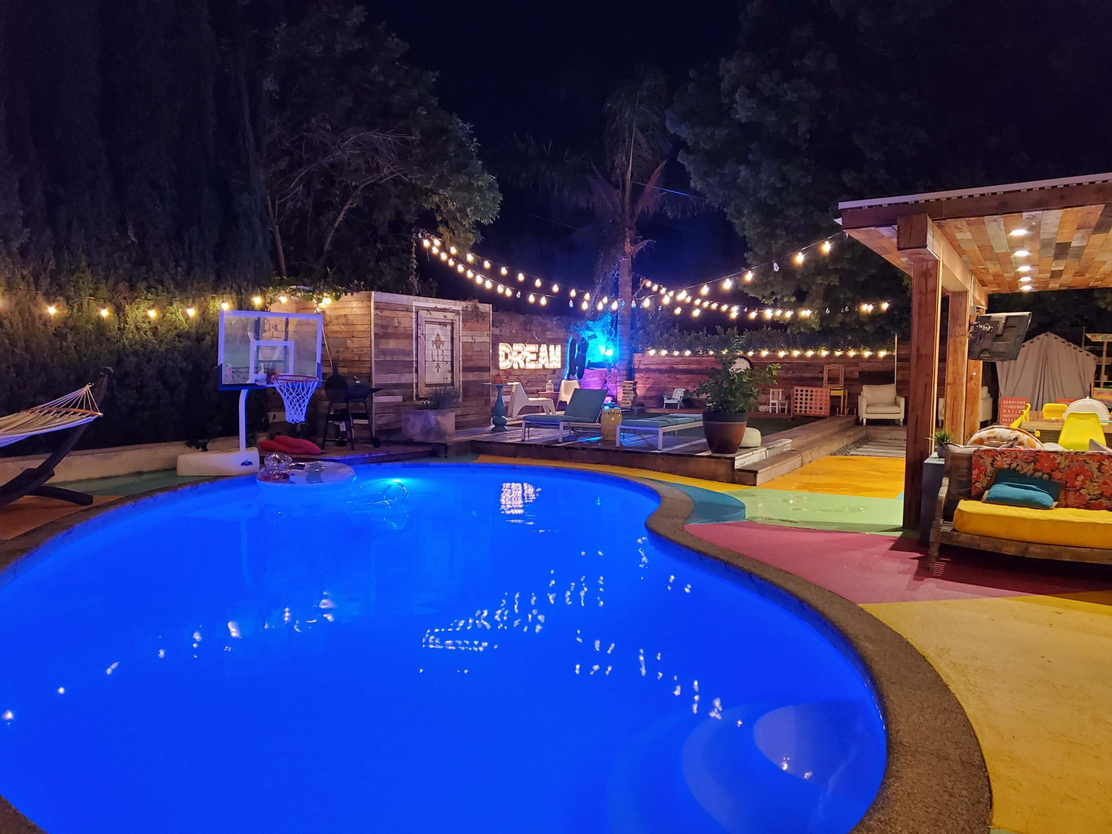 The scene features a brightly lit pool area at night, surrounded by lounge chairs and decorative string lights under a canopy of trees.