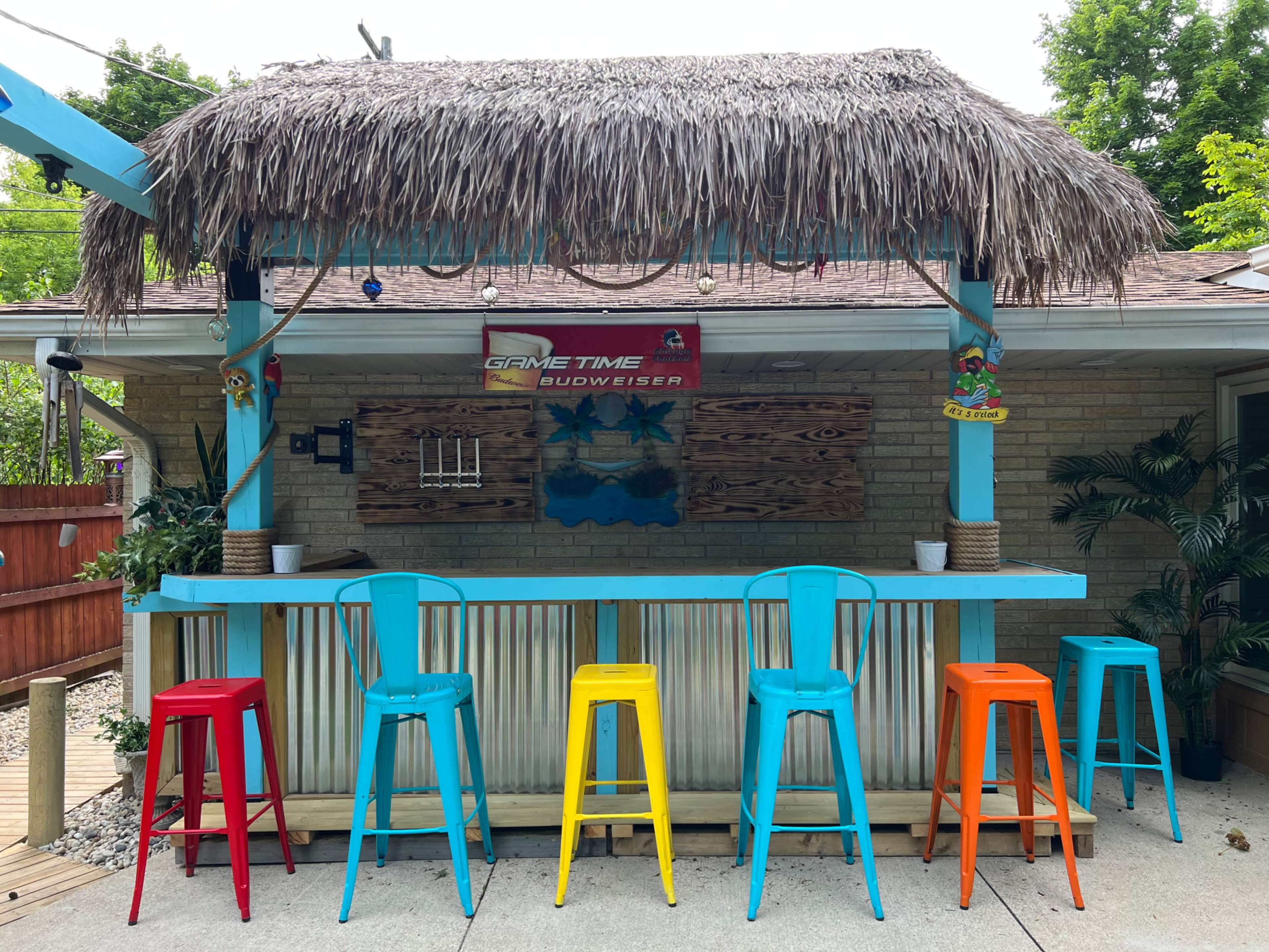 A thatched-roof outdoor bar features brightly colored metal stools arranged in front of a wooden counter.