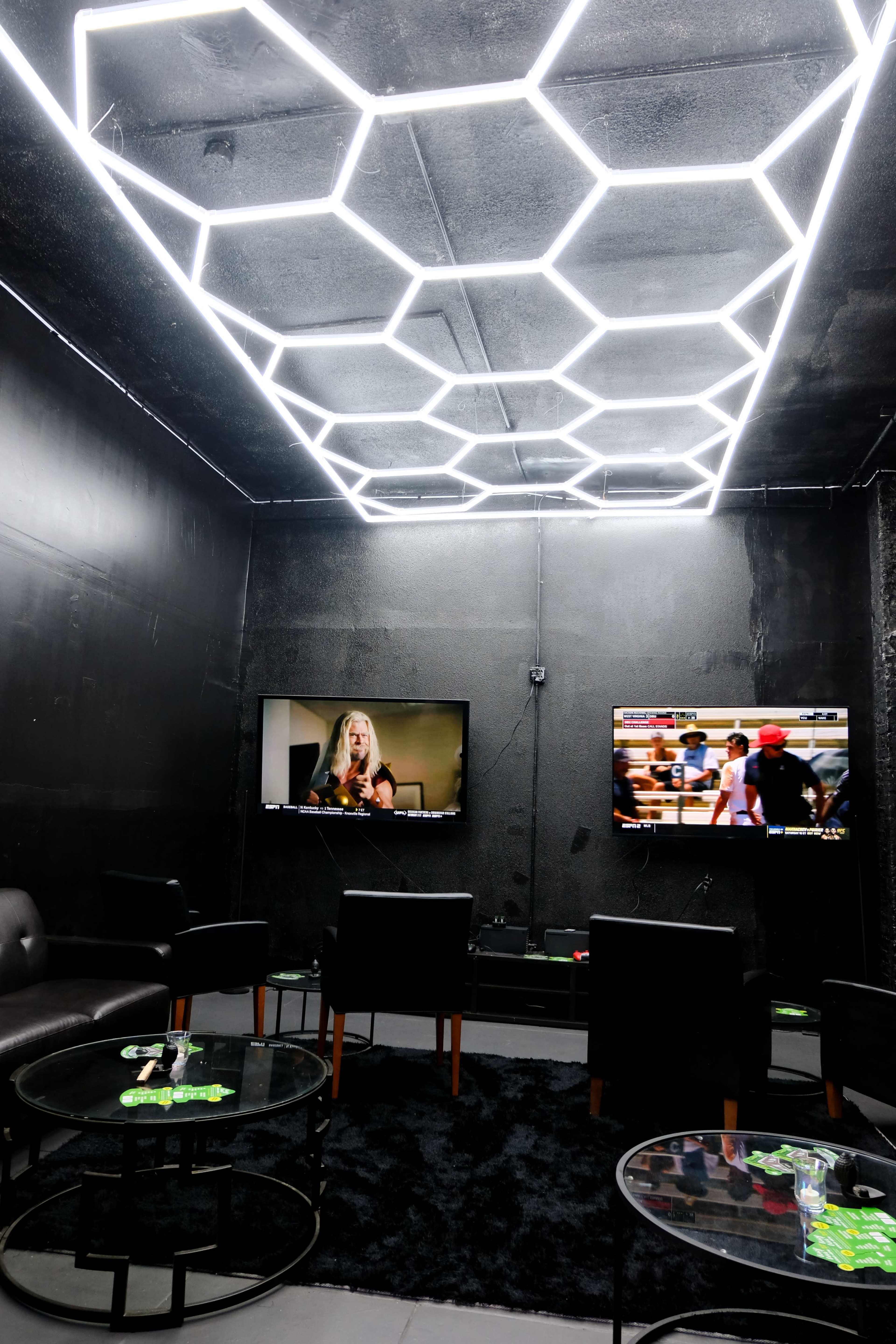 A modern lounge with a hexagonal neon ceiling light, two televisions on the walls, and black furniture arranged around glass tables.