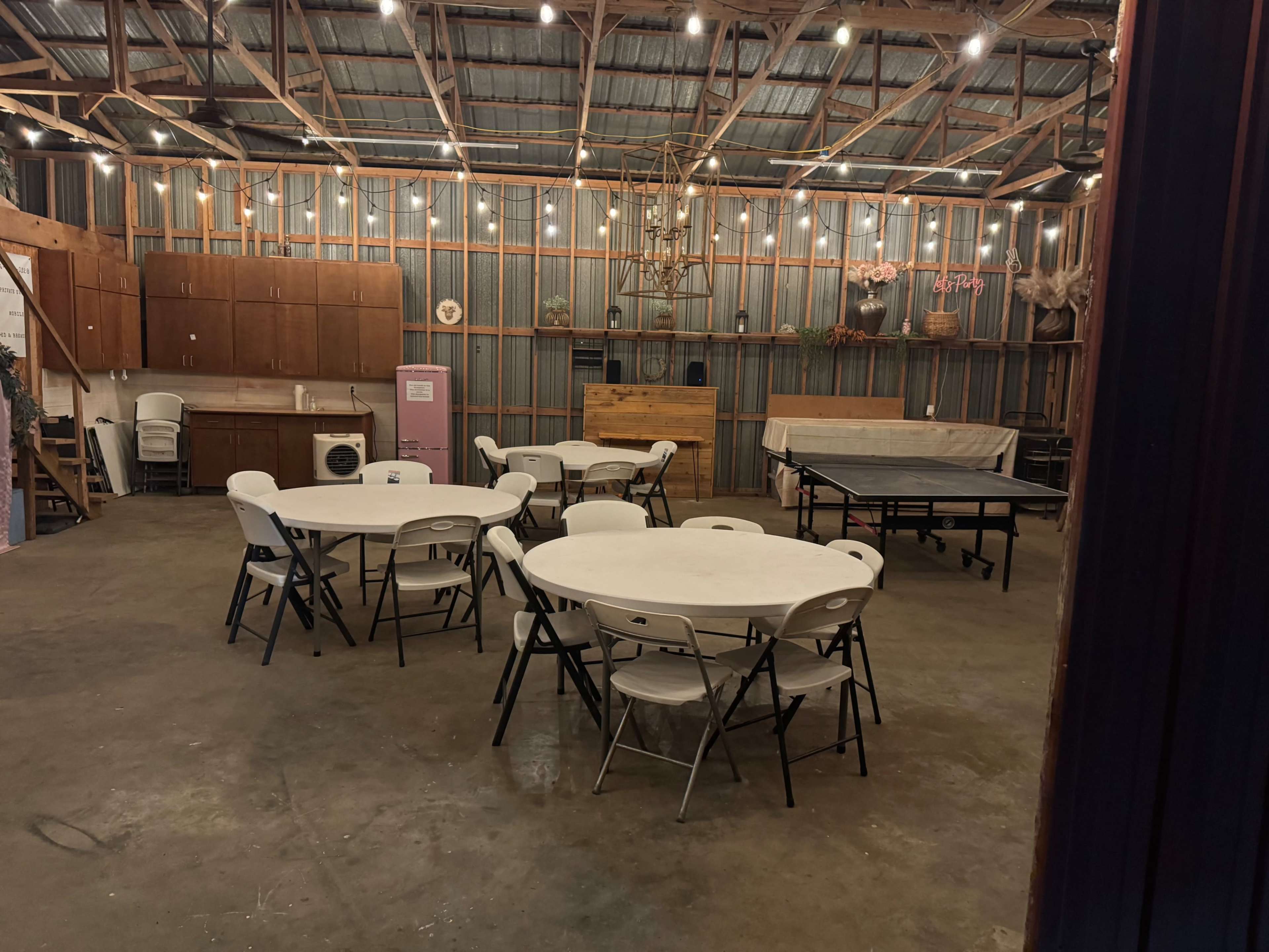 The interior of a barn-style venue features several round tables with white chairs, string lights overhead, and a wooden stage area at the front.