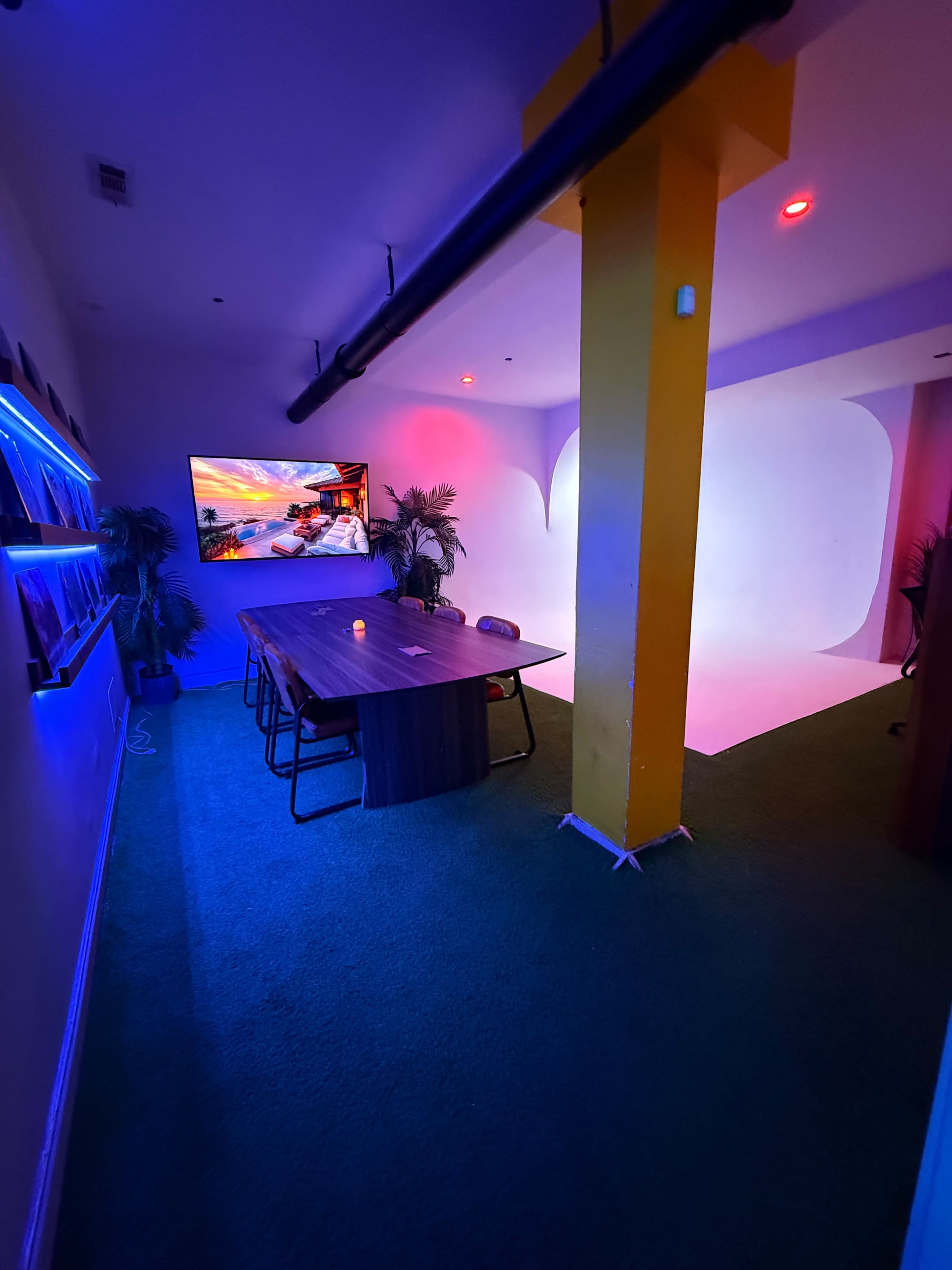 The image shows a modern meeting room with a large table, a colorful wall-mounted screen, and vibrant lighting.