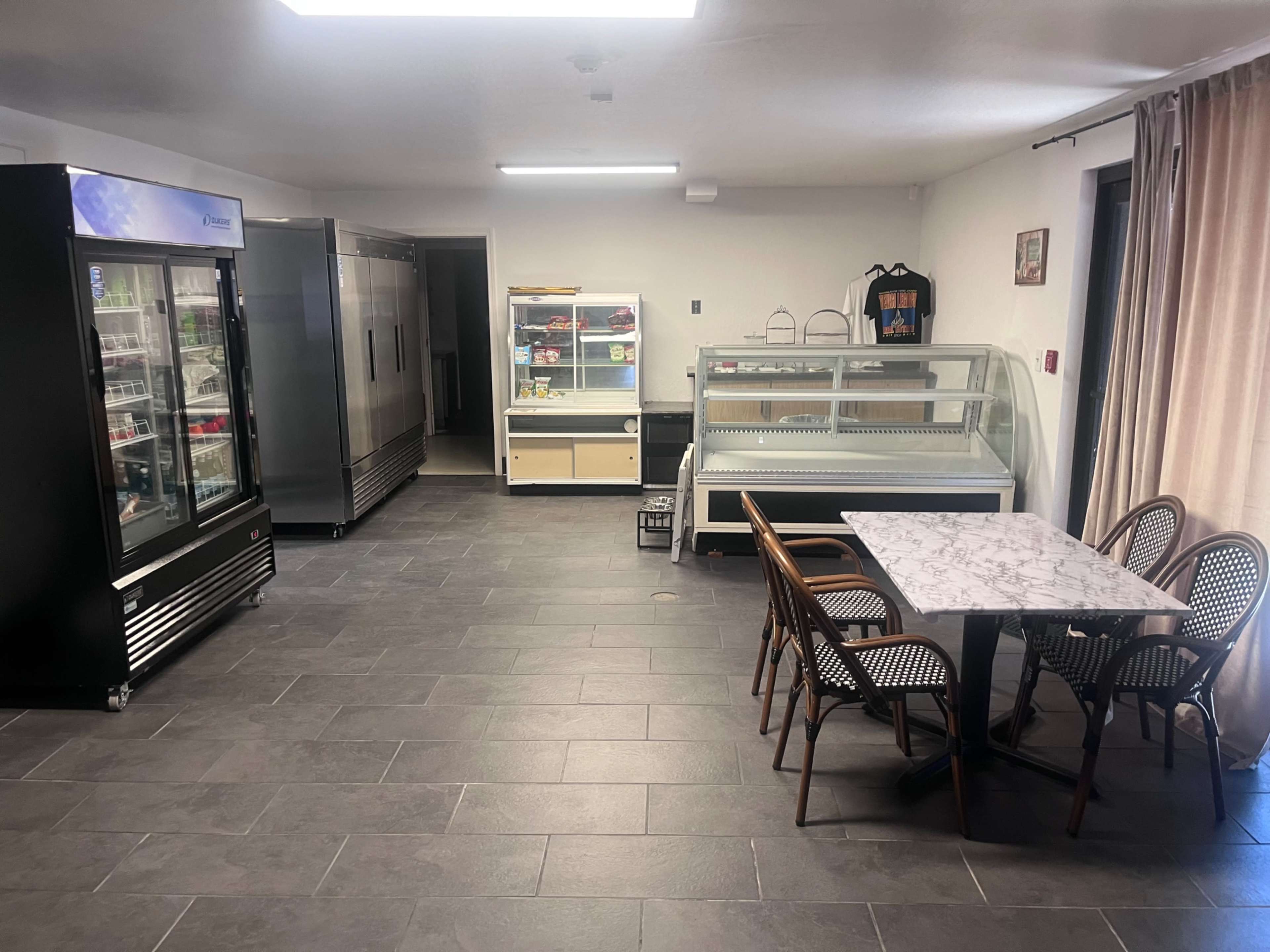 The image shows a spacious interior of a commercial kitchen or café featuring a display case, two large refrigerators, and a dining area with a marble-top table and wicker chairs.