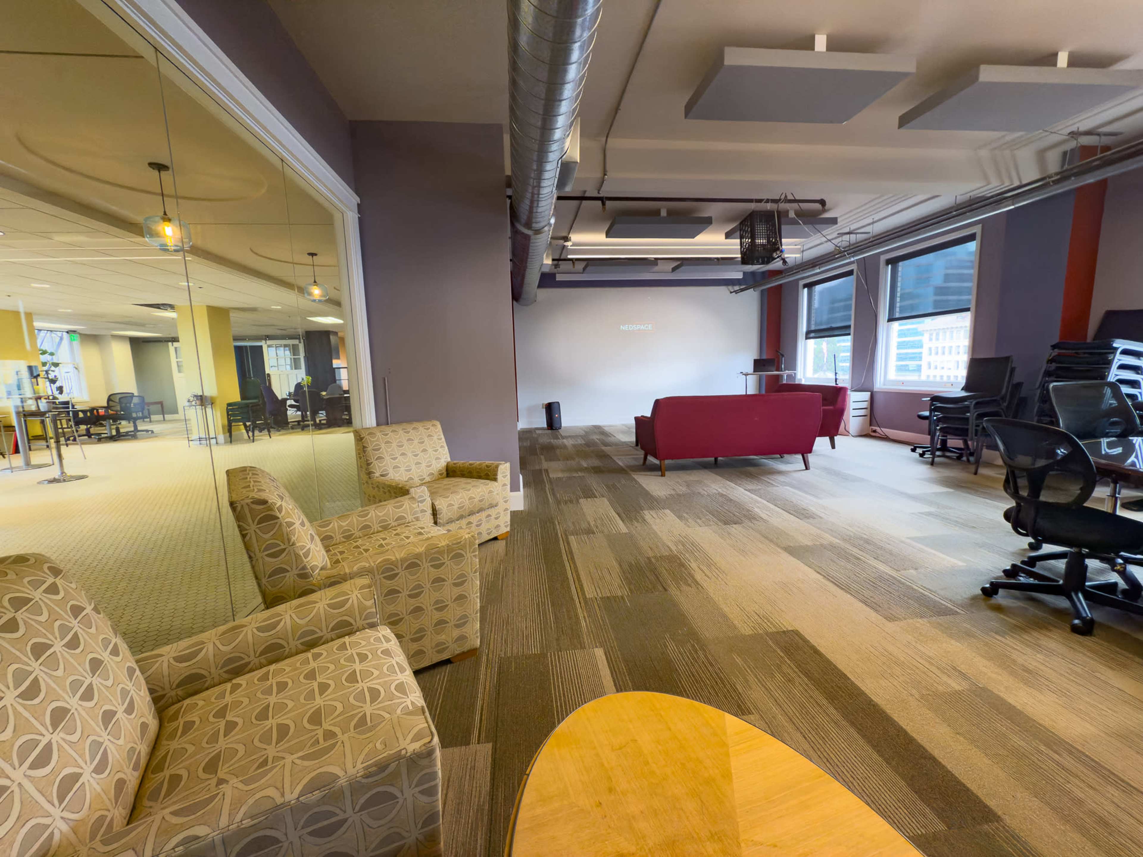 The image shows a modern office space with a seating area featuring patterned chairs and a table, along with unoccupied workstations and a red sofa in the background.