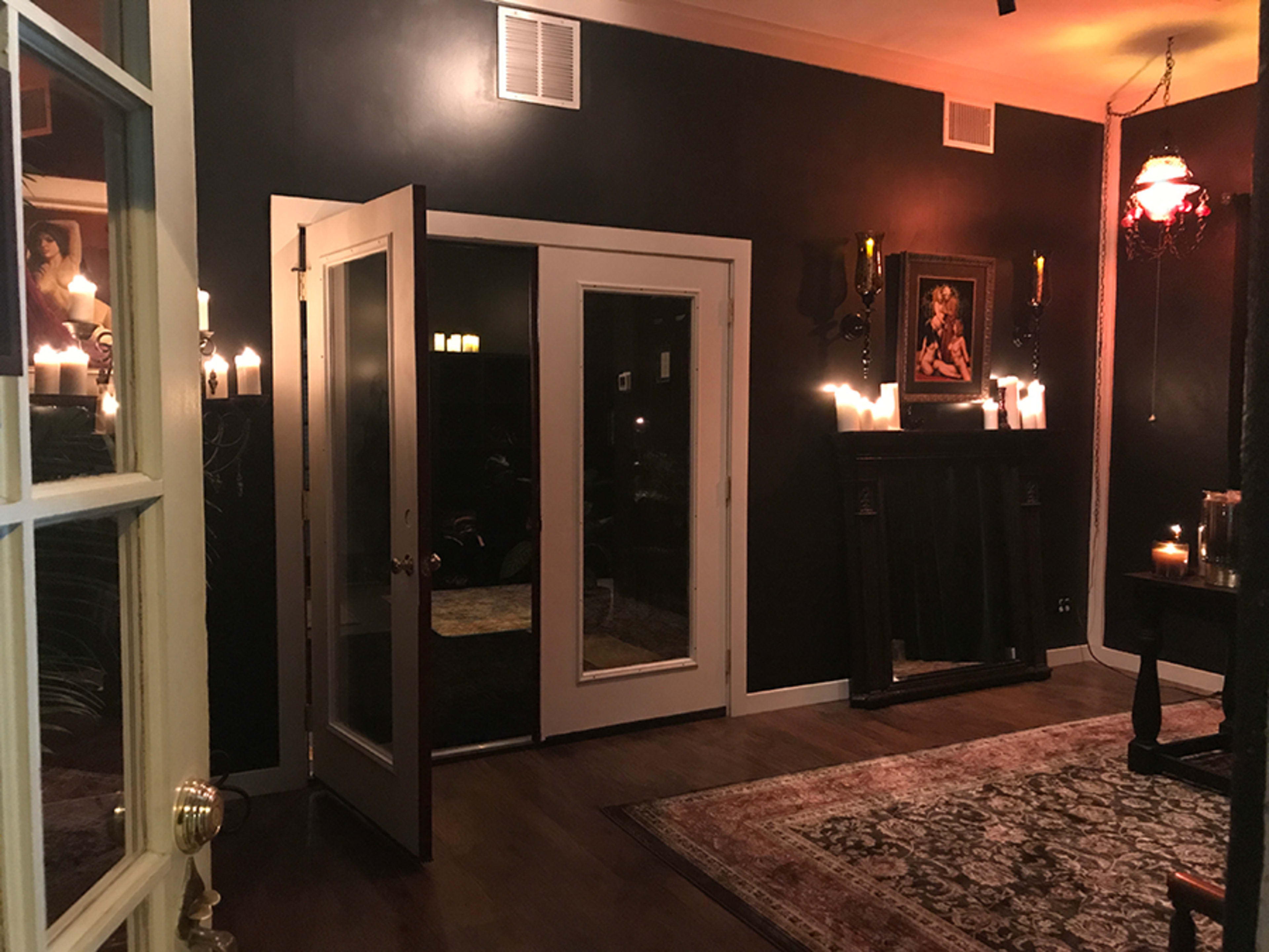 A dimly lit room features a pair of open double doors, a dark-painted wall, a fireplace with candles, and a large rug on the wooden floor.